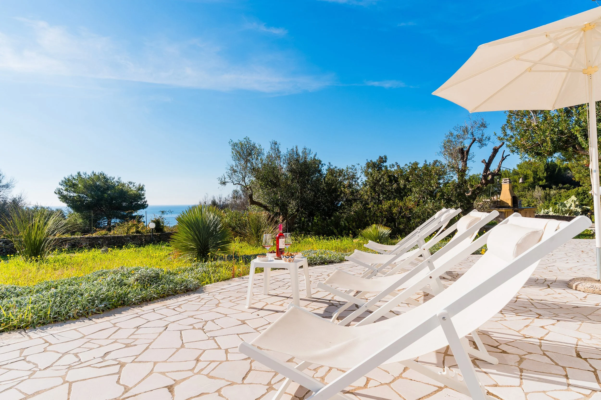 A sunny outdoor patio with white lounge chairs, a table with snacks and drinks, and a large white umbrella. There are trees and bushes, and a view of the ocean in the background.