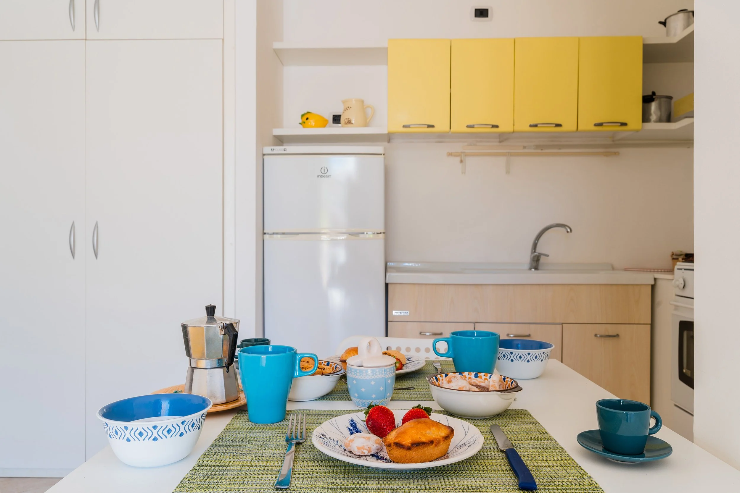 A breakfast table set with blue cups, plates, and a variety of pastries, strawberries, and a cooked egg, in a kitchen with white and yellow cabinets.