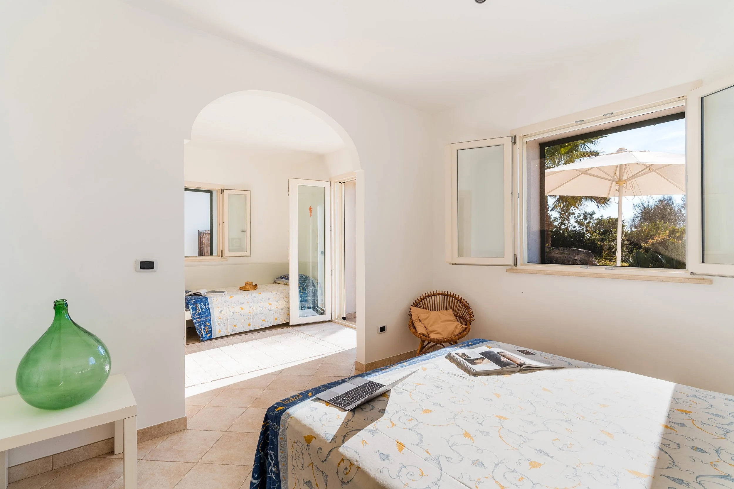 A bright bedroom with an open window showing a balcony with a large umbrella and trees outside, a table with a patterned tablecloth, an open laptop, a small basket chair with a cushion, and a glimpse of a bed with a blue and white bedspread.