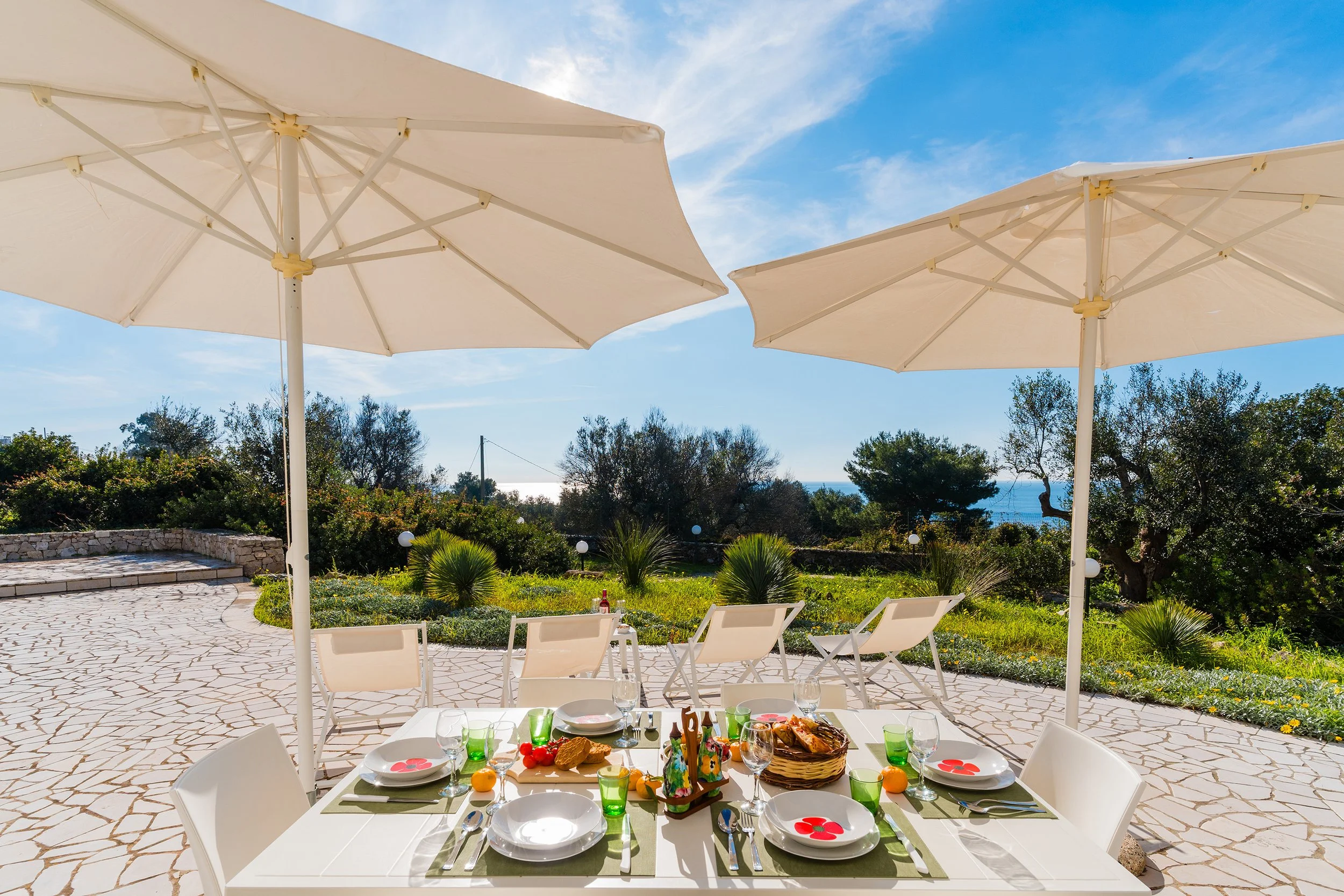 Outdoor dining setup with a table, chairs, and umbrellas in a garden or patio area on a sunny day.