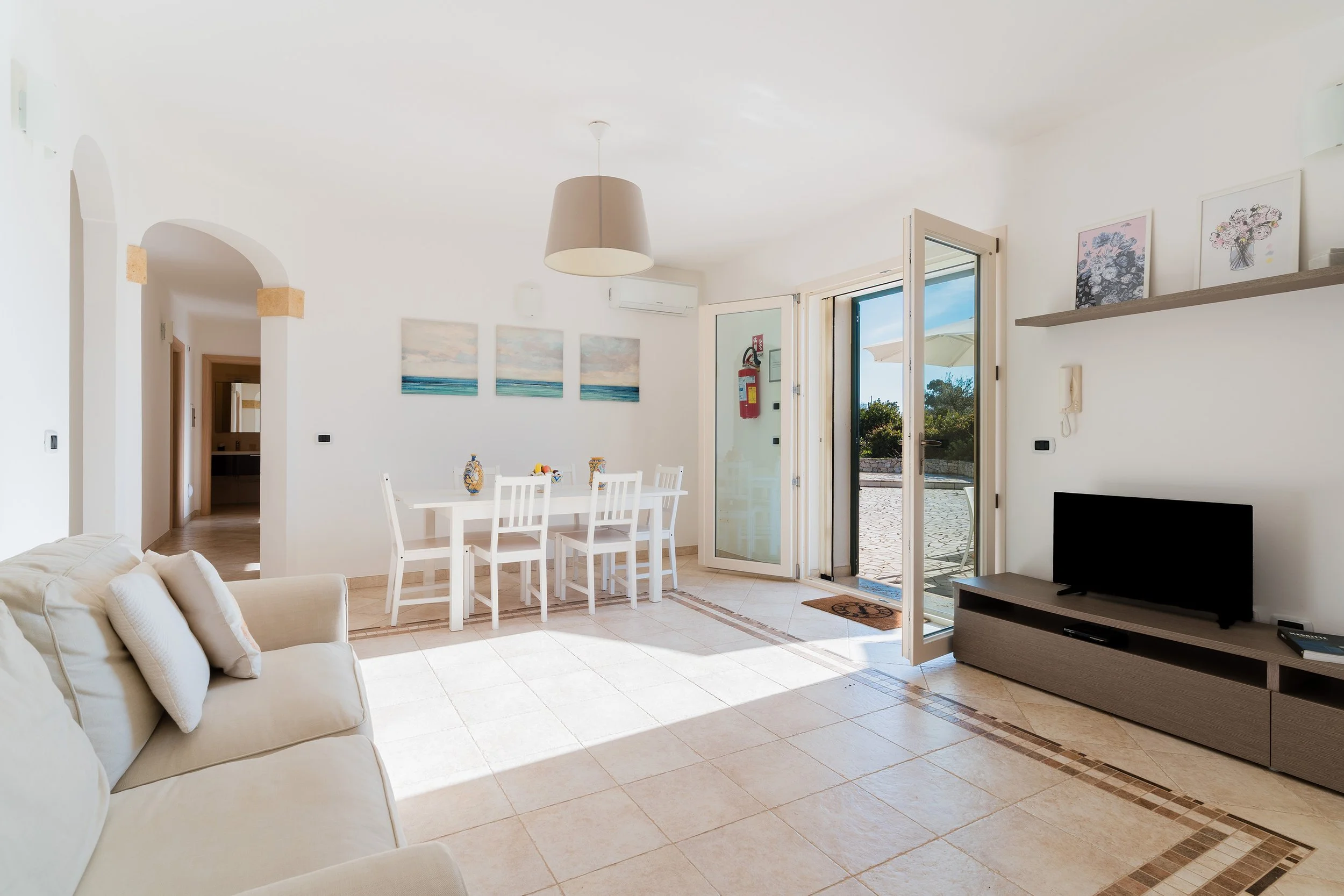 A bright, airy living and dining area with white walls, tiled floors, and open double doors leading to an outdoor patio, decorated with paintings, a beige sofa, a white dining table with chairs, and a modern TV stand.
