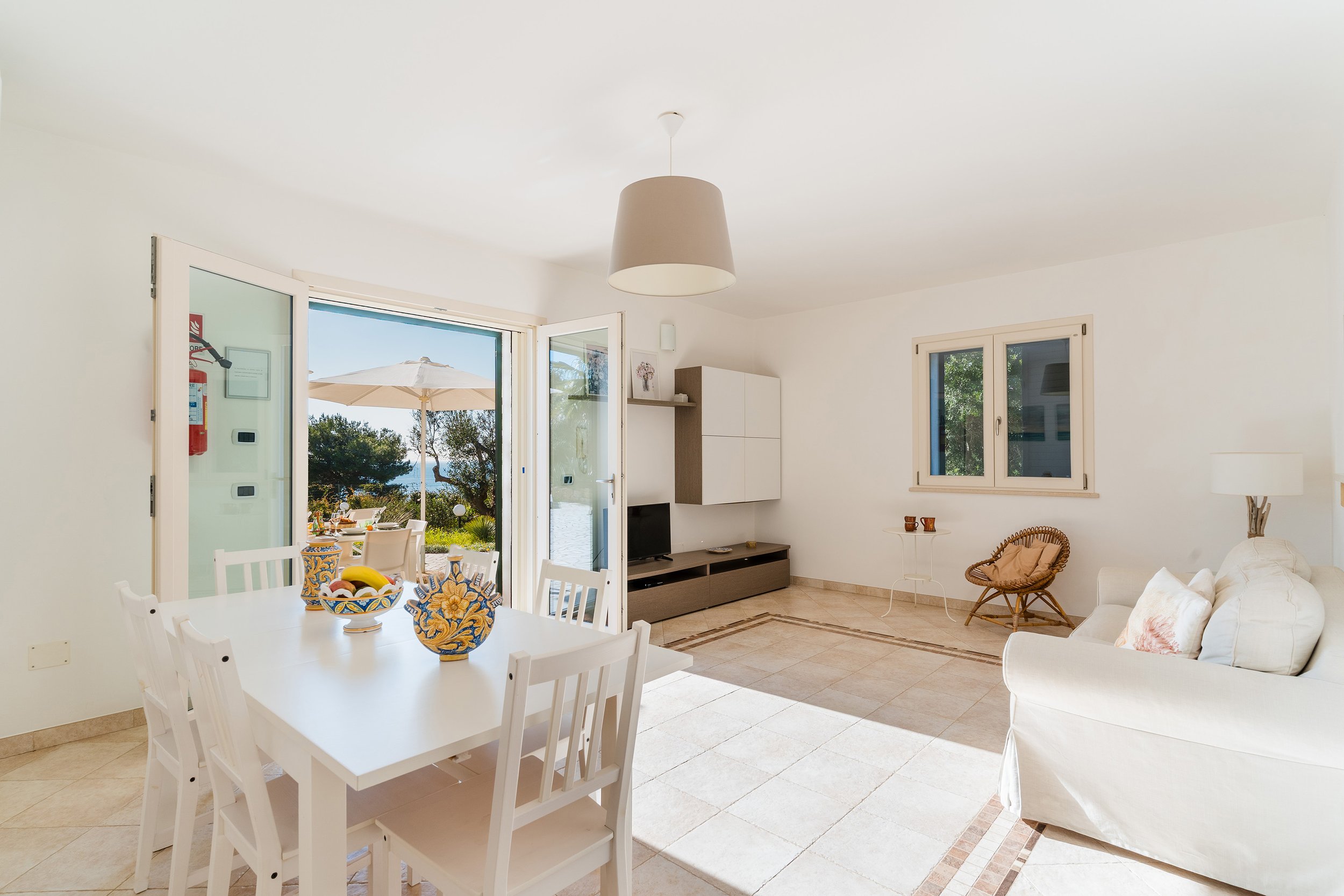 Bright living room with white walls, open glass door leading to outdoor patio, and views of trees and water, featuring a dining table with colorful vases, a television, a sofa, and wicker chairs.