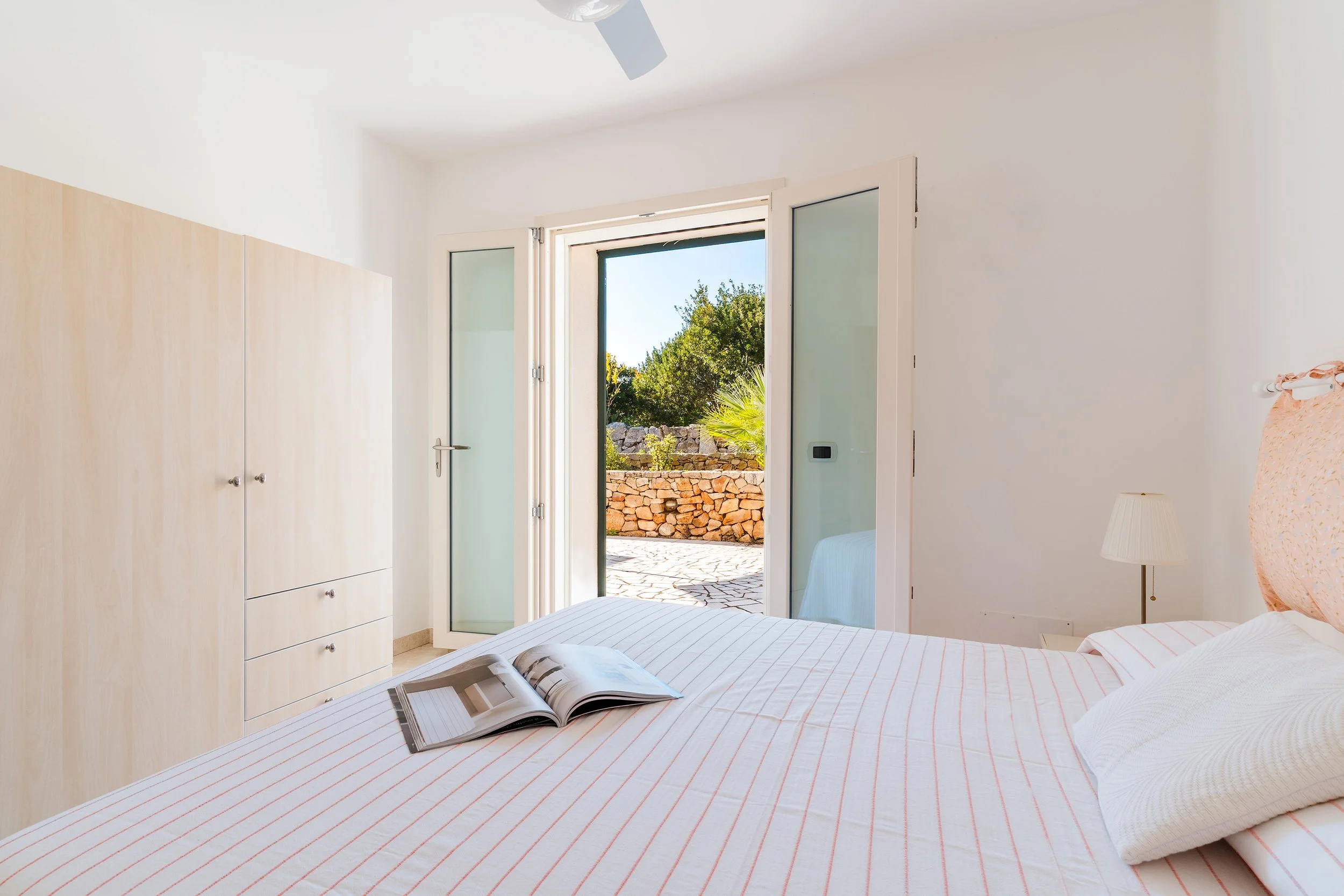A bedroom with a bed, an open magazine, a white wardrobe, a bedside lamp, and an open door leading outside to trees and a stone wall.