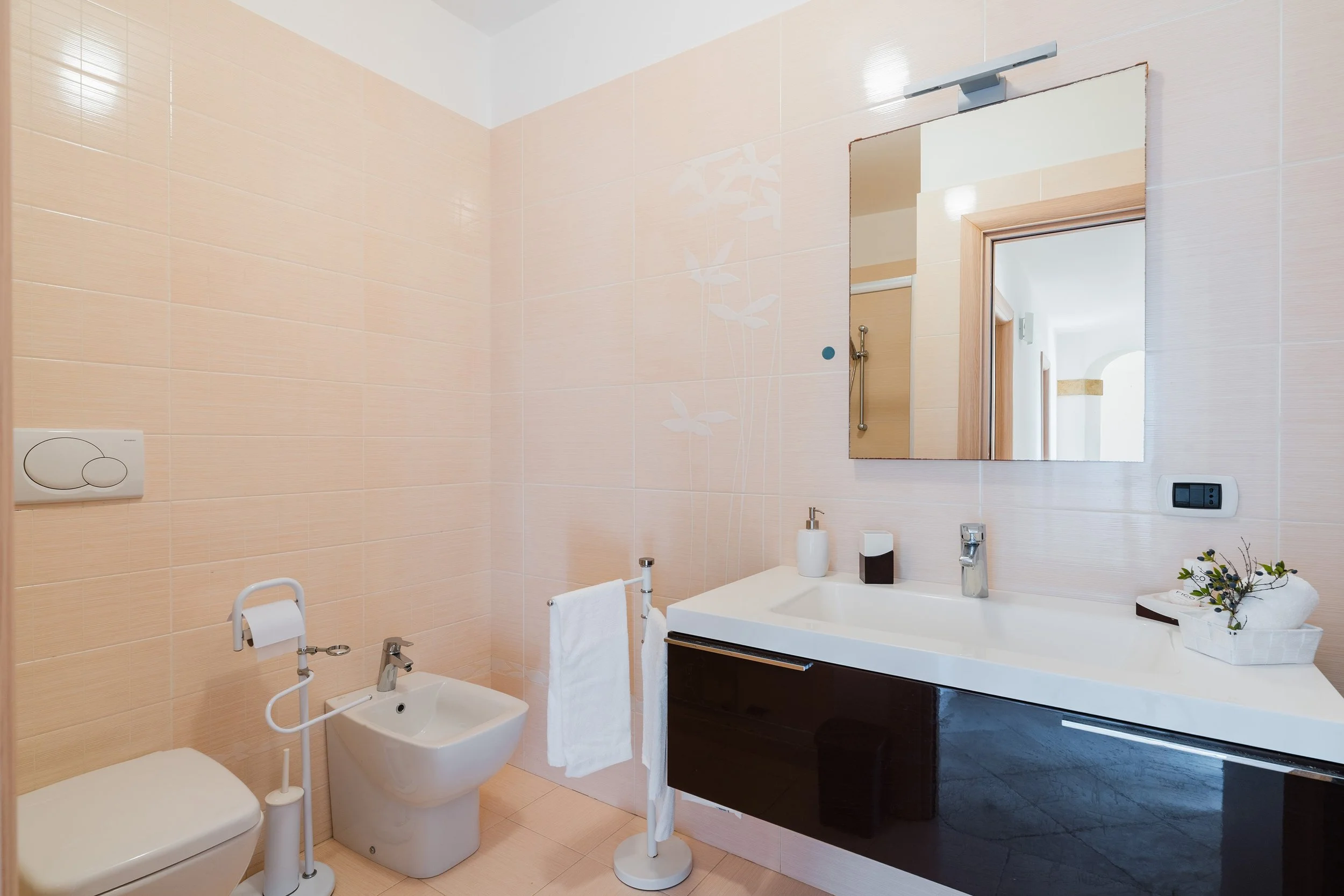 Bathroom with beige tiled walls, a mirror above a sink, a toilet, a bidet, and a small potted plant on the countertop.