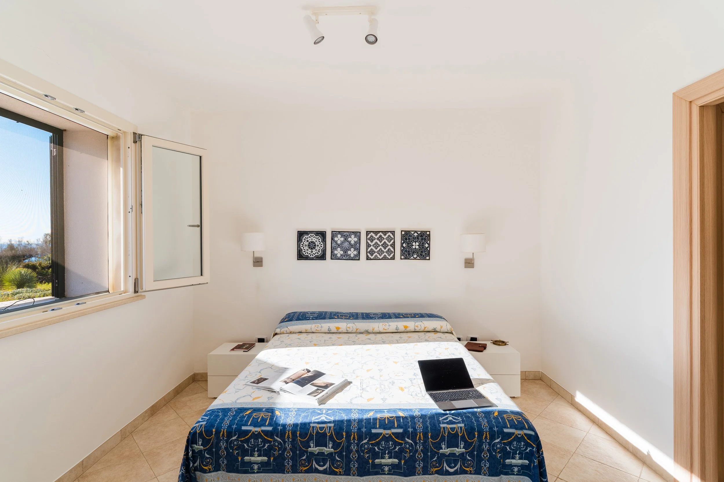 A hotel room with a bed, matching nightstands, white walls, and decorative tiles above the bed. The room has a window with an open shutter showing outdoor scenery and a wooden doorframe on the right. There are some magazines and a laptop on the bed.