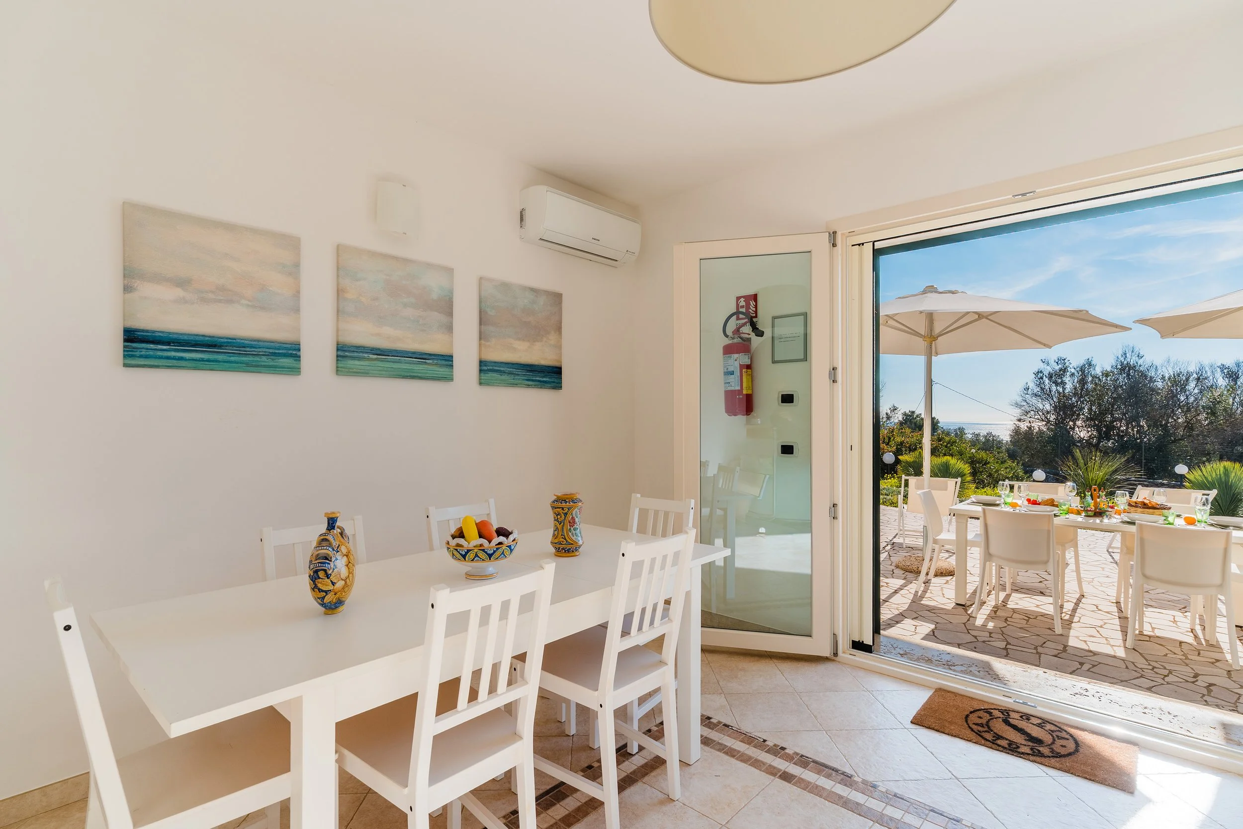 Dining room with white table and chairs, decorative vases, and a bowl of fruit, opening to outdoor patio with table, chairs, umbrellas, and a view of trees and blue sky.
