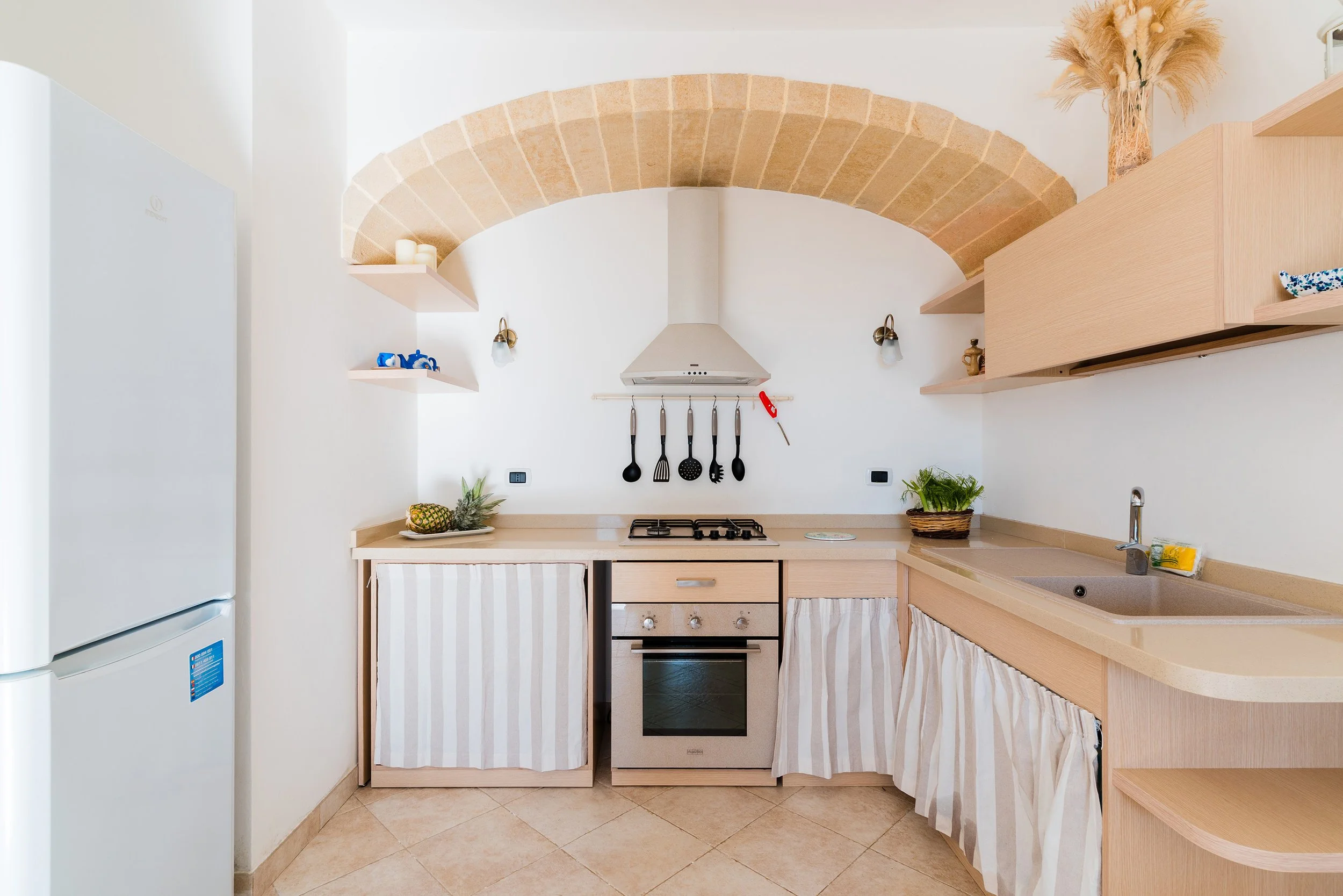Kitchen with beige countertops, striped curtains, and open wooden shelves, featuring a pineapple, potted plants, and kitchen utensils hanging above the stove.