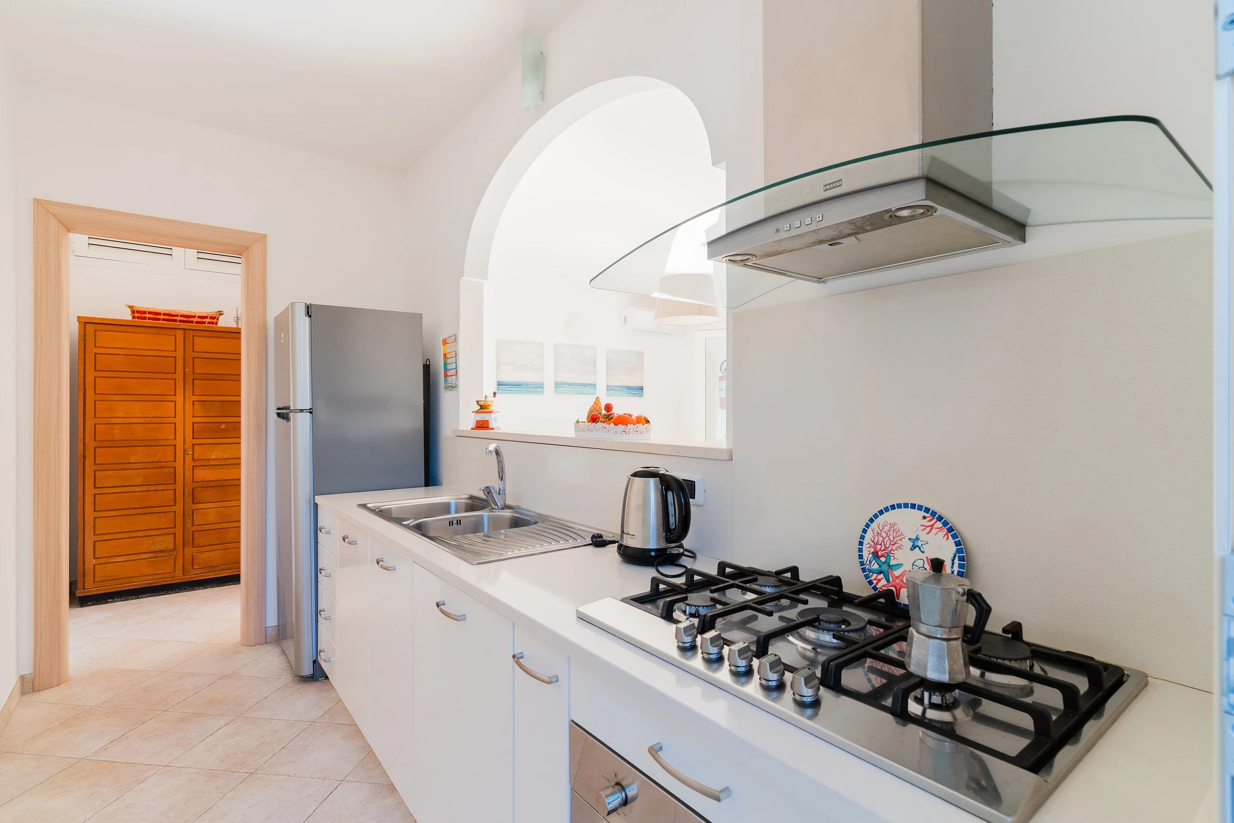 Kitchen with white cabinets, stainless steel sink, electric kettle, gas stove, and a decorative plate with fish and coral on the wall. Bright space with natural light, wooden door to a storage area, and a view into another room with paintings and a f