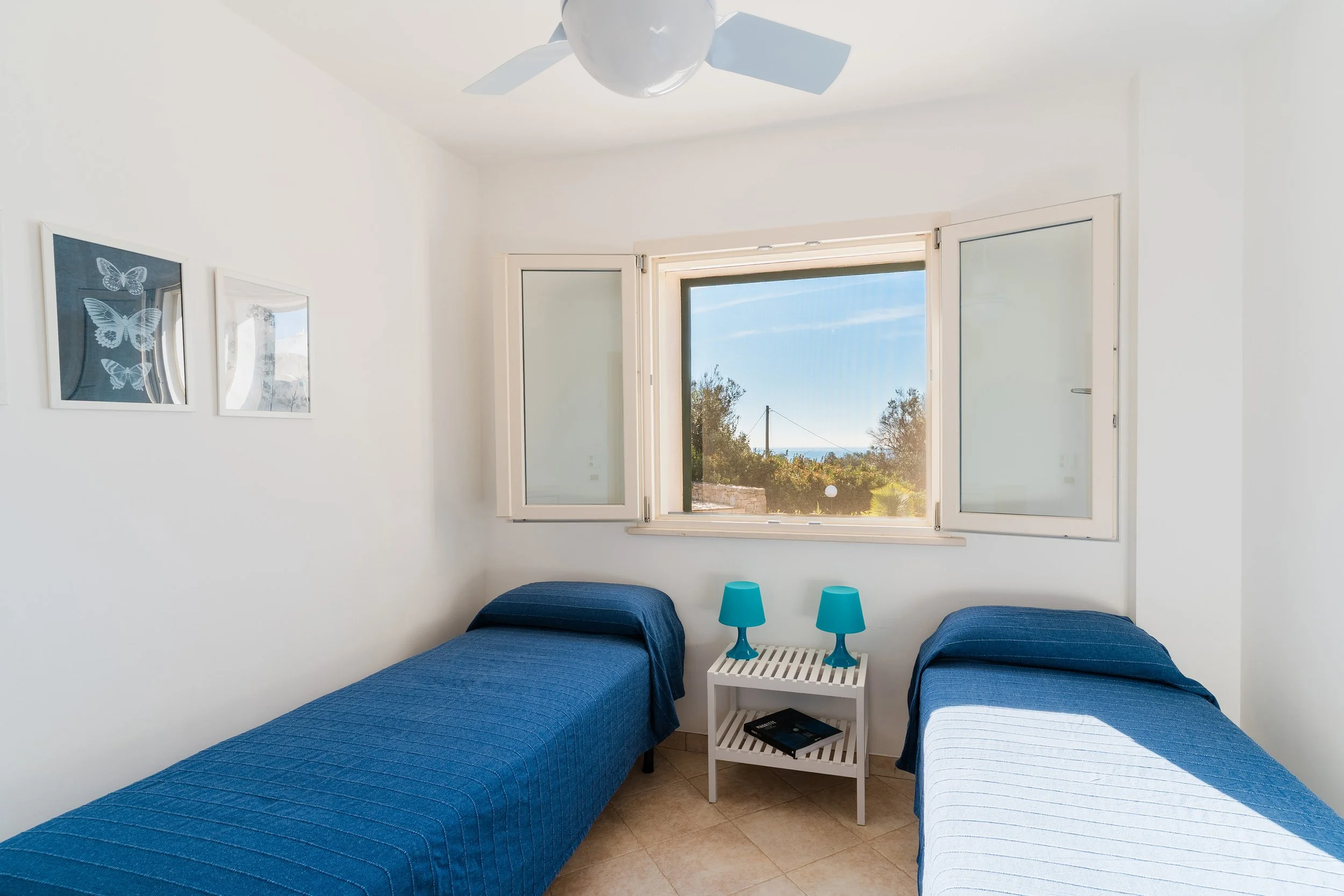 A bright bedroom with a large open window showing trees and the sky outside. There are two twin beds with blue bedspreads, a white bedside table with two turquoise lamps, and three framed pictures of butterflies on the wall.