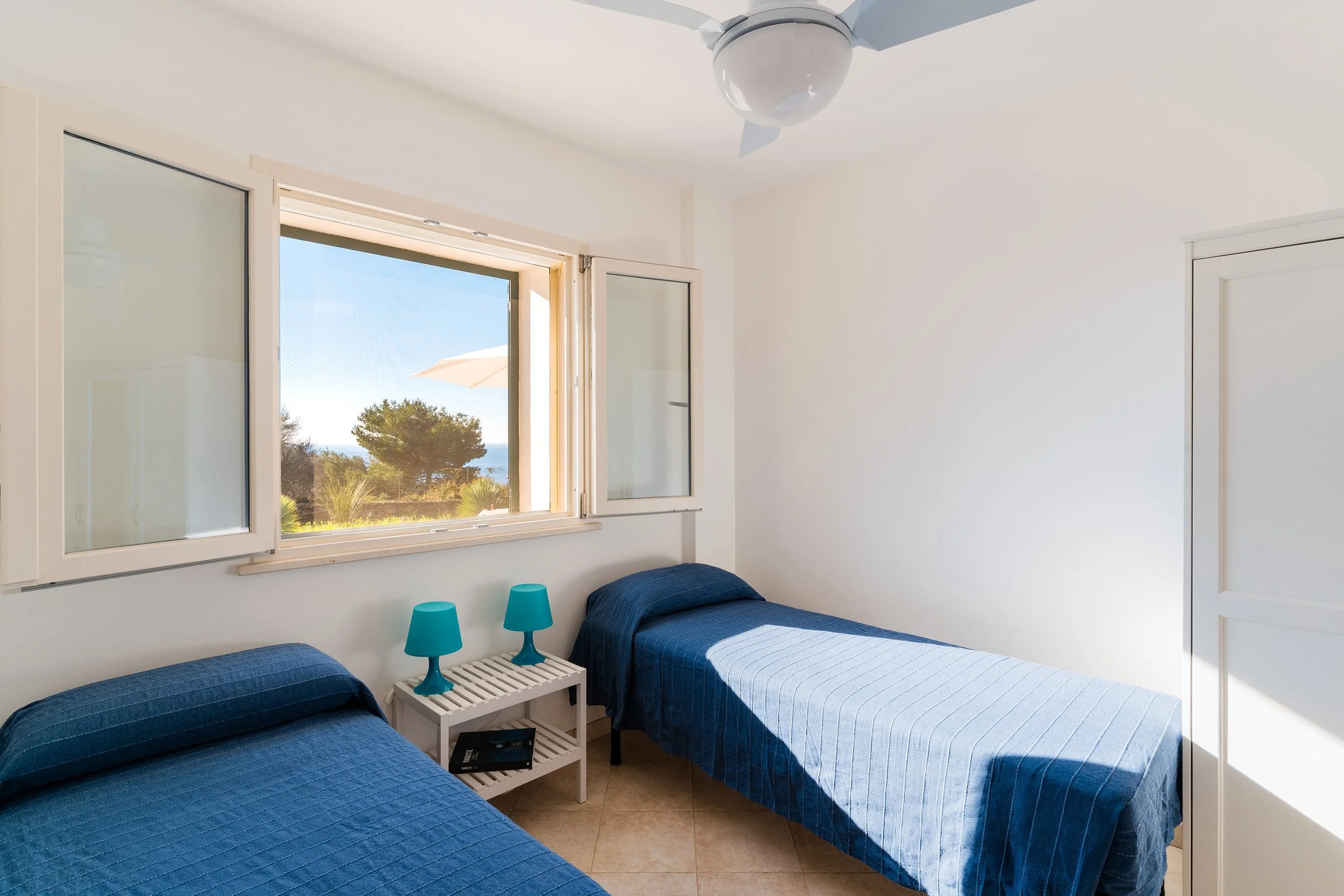 A bedroom with two twin beds covered with blue quilted bedspreads. Between the beds, there is a small white slatted nightstand with two turquoise table lamps and a black telephone. The room has a large window with an open window shutter, showing outd