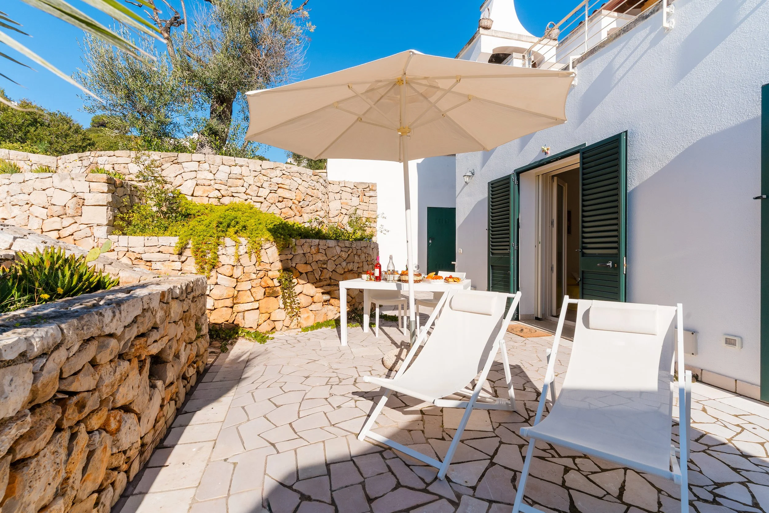 A sunny outdoor patio with a large beige umbrella, white chairs, a small table with drinks and snacks, stone walls, and a white building with green shutters and open doors.