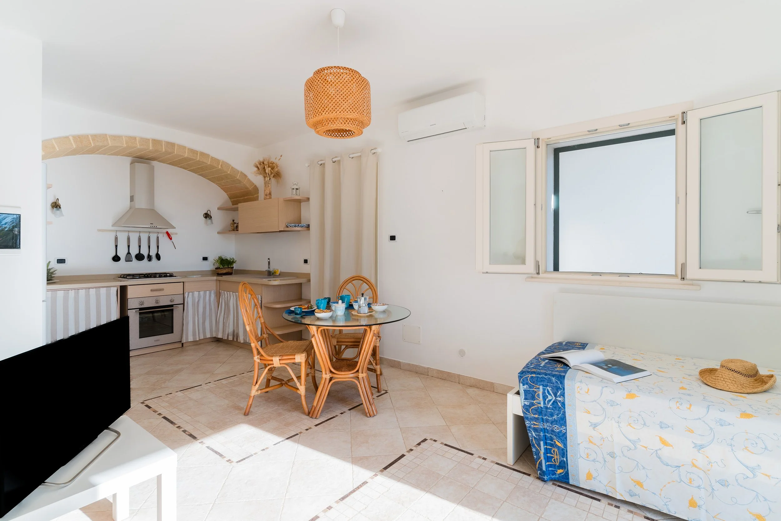 A bright, white room with a small kitchen area featuring a stove, sink, and beige cabinetry. There is a round dining table with four wooden chairs, set with breakfast dishes. A bed with blue and yellow linens and an open book is visible in the corner