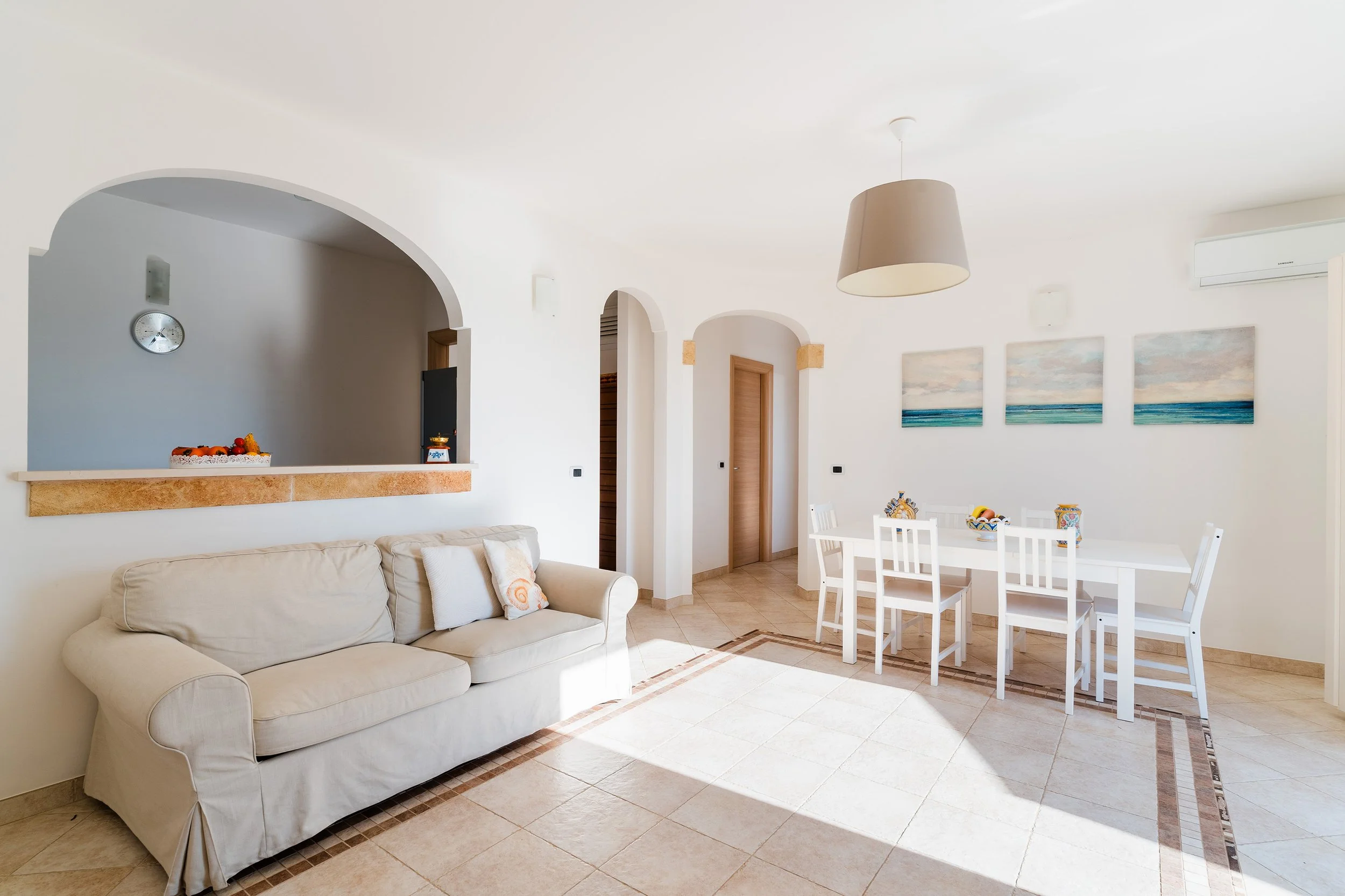 Bright living room with white walls, beige sofa, and a white dining table with chairs. Decor includes three seascape paintings, a ceiling lamp, wall-mounted air conditioning, and a clock.