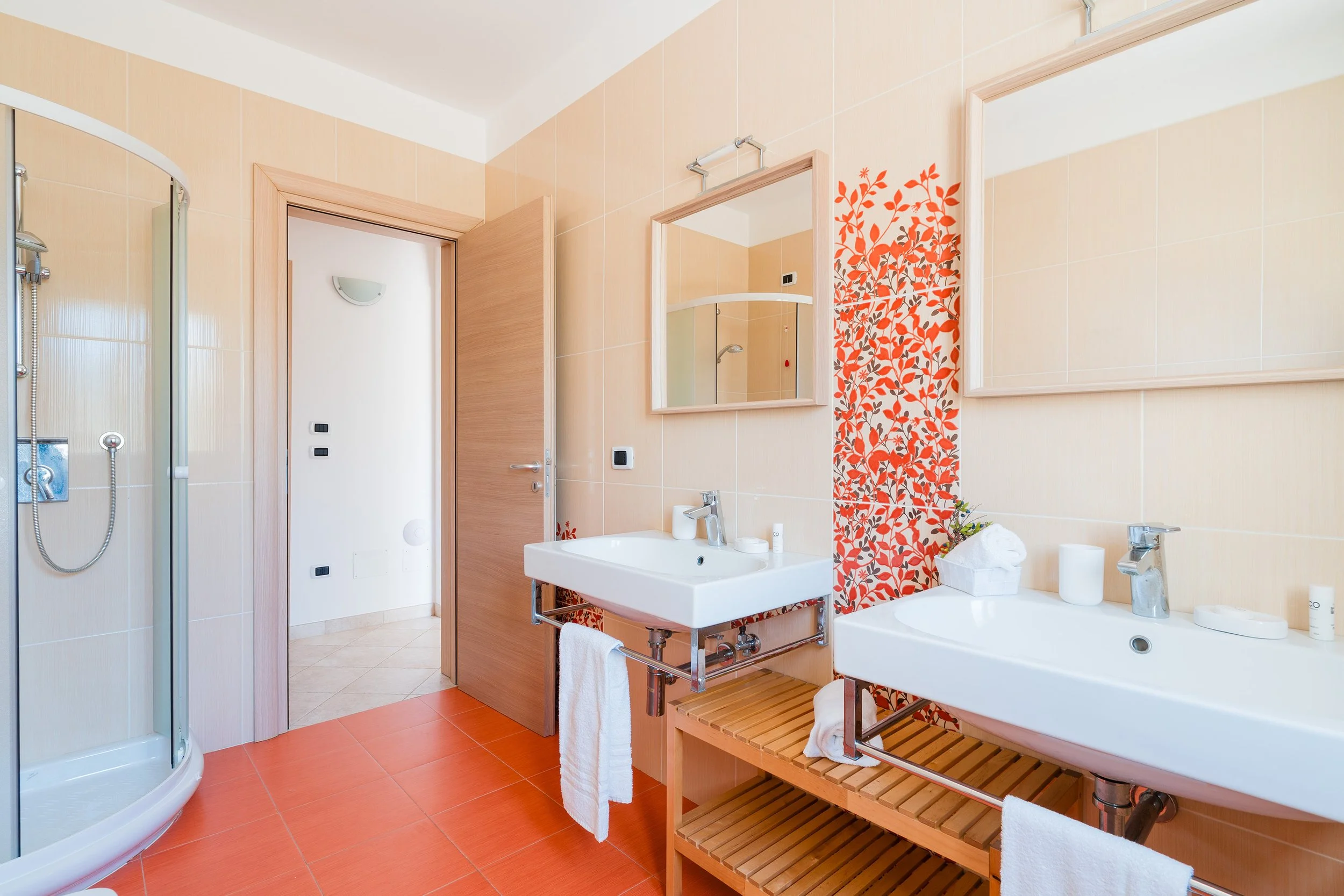 A modern bathroom with beige tiled walls and orange floor tiles. It features a curved glass shower enclosure on the left, two white pedestal sinks with matching mirrors above, and a wooden shelf beneath one sink. A decorative strip with orange and gr