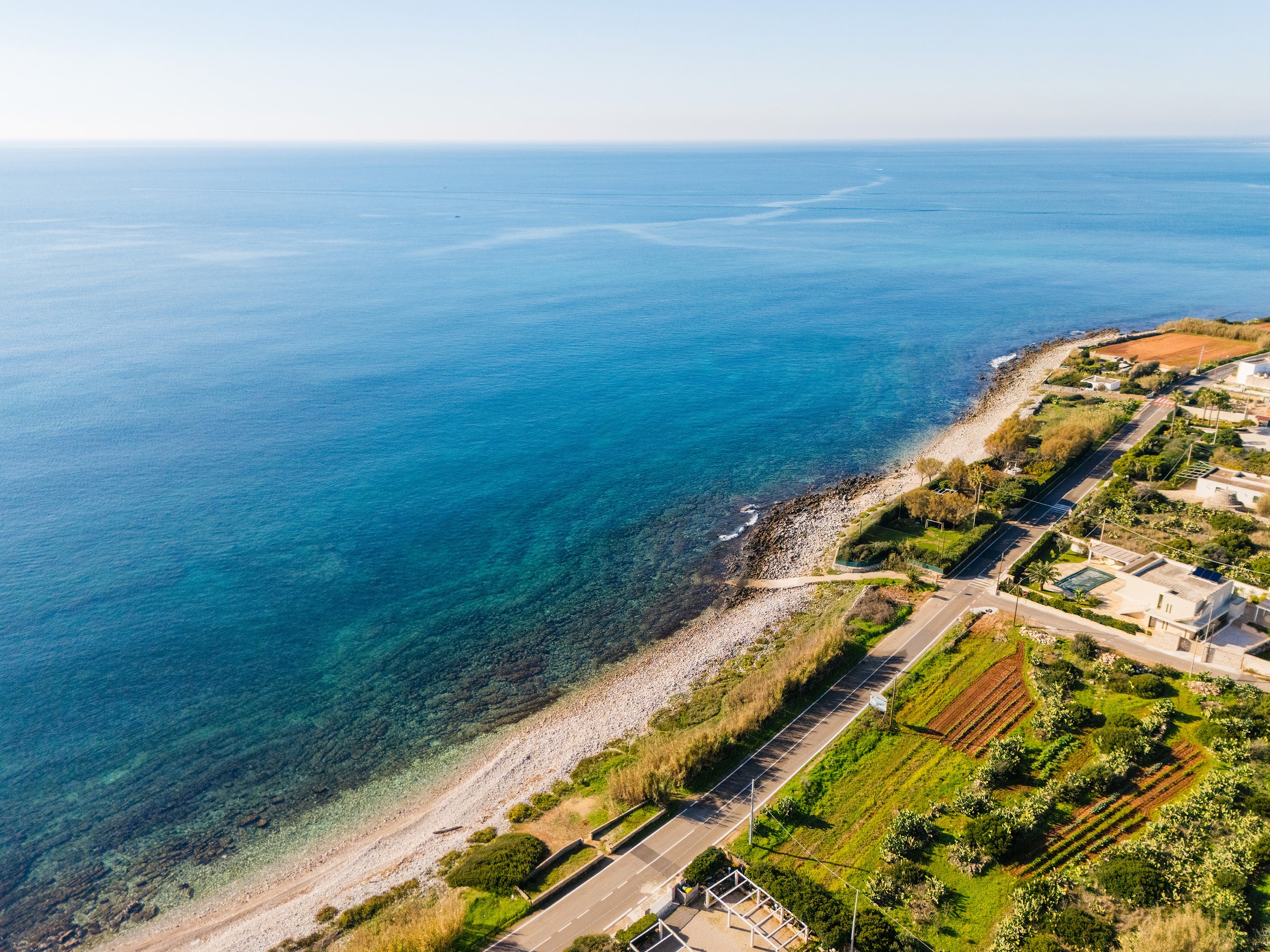 Aerial view of a coastline with clear blue water, a rocky beach, and residential houses with green trees and gardens along a road.