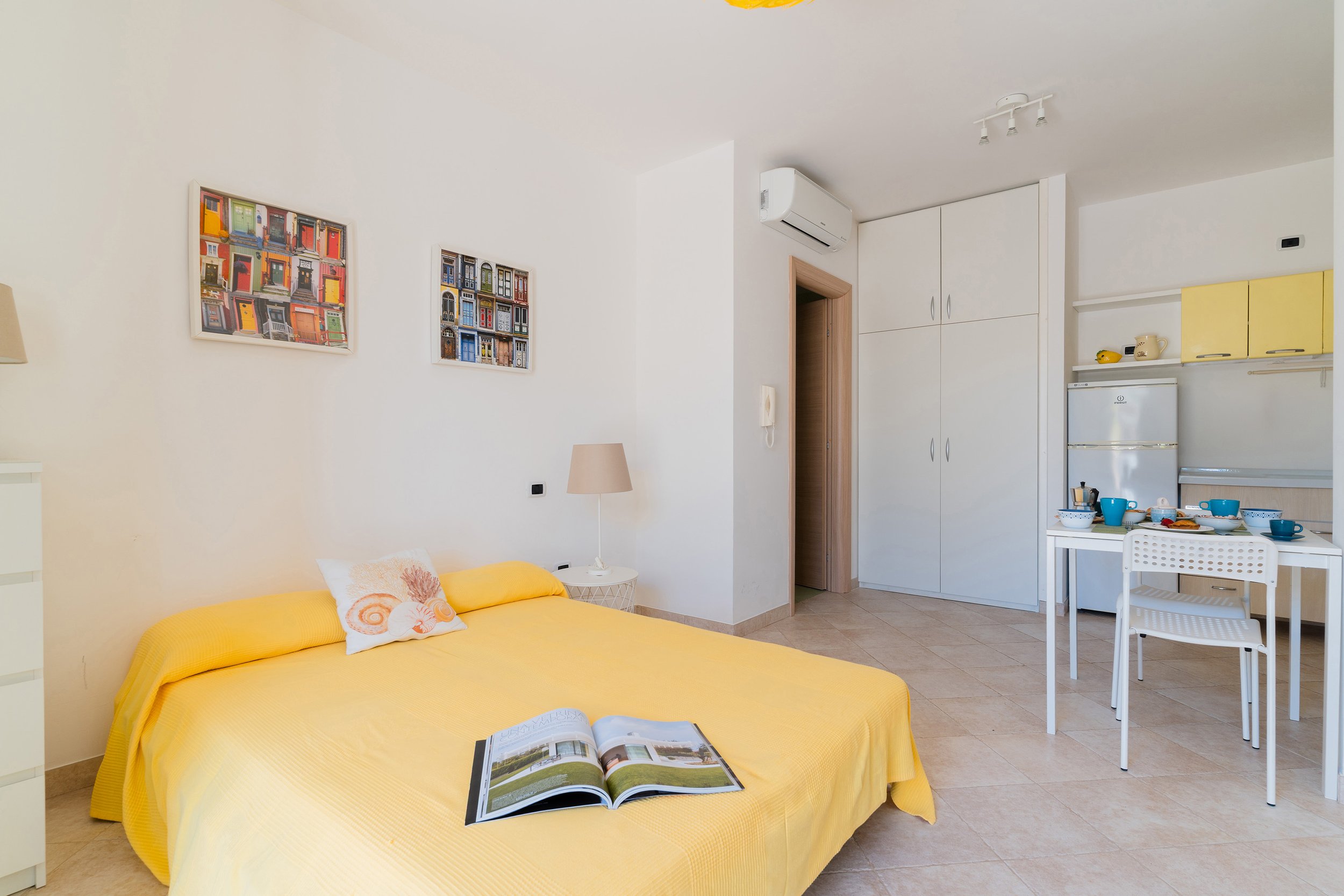 A cozy bedroom with a yellow bedspread, an open magazine, and wall art of colorful doors, near a kitchenette with white cabinets and a small dining table with blue dishes.