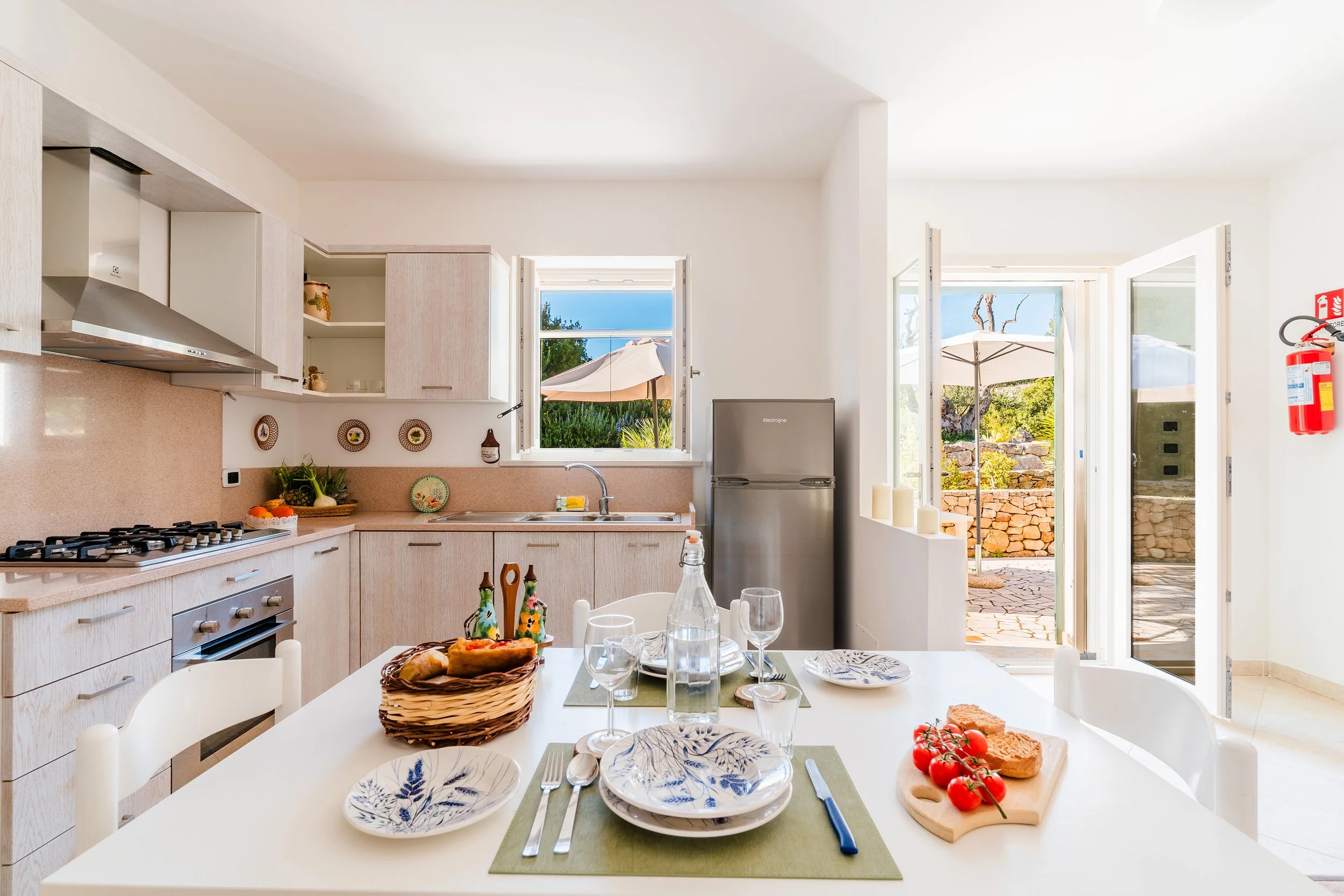 A bright, modern kitchen with open windows showing outdoor patio and garden. There is a dining table with place settings, a basket of bread, and fresh tomatoes. Food items and tableware are arranged on the counter and table, with a refrigerator and kitchen cabinets in the background.
