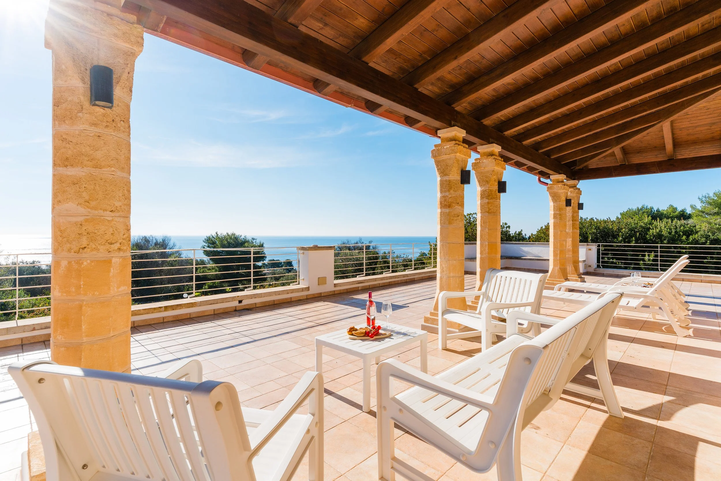 A spacious outdoor terrace with white lounge chairs, a small side table with a plate of snacks and drinks, under a wooden pergola with stone columns. The background shows a clear blue sky, ocean, and greenery.