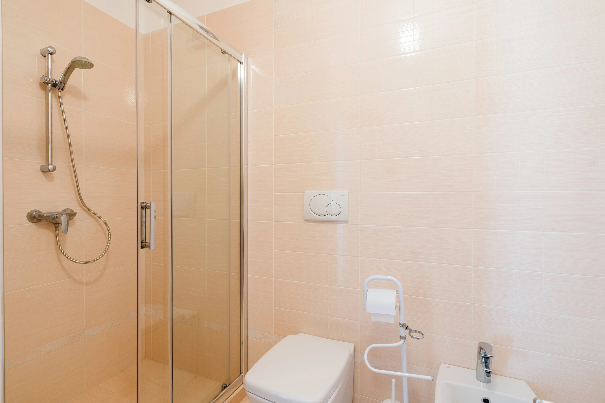 A small bathroom with beige tiled walls, a glass shower enclosure with a silver frame, a handheld showerhead, a toilet, a bidet, and a sink with a silver faucet.