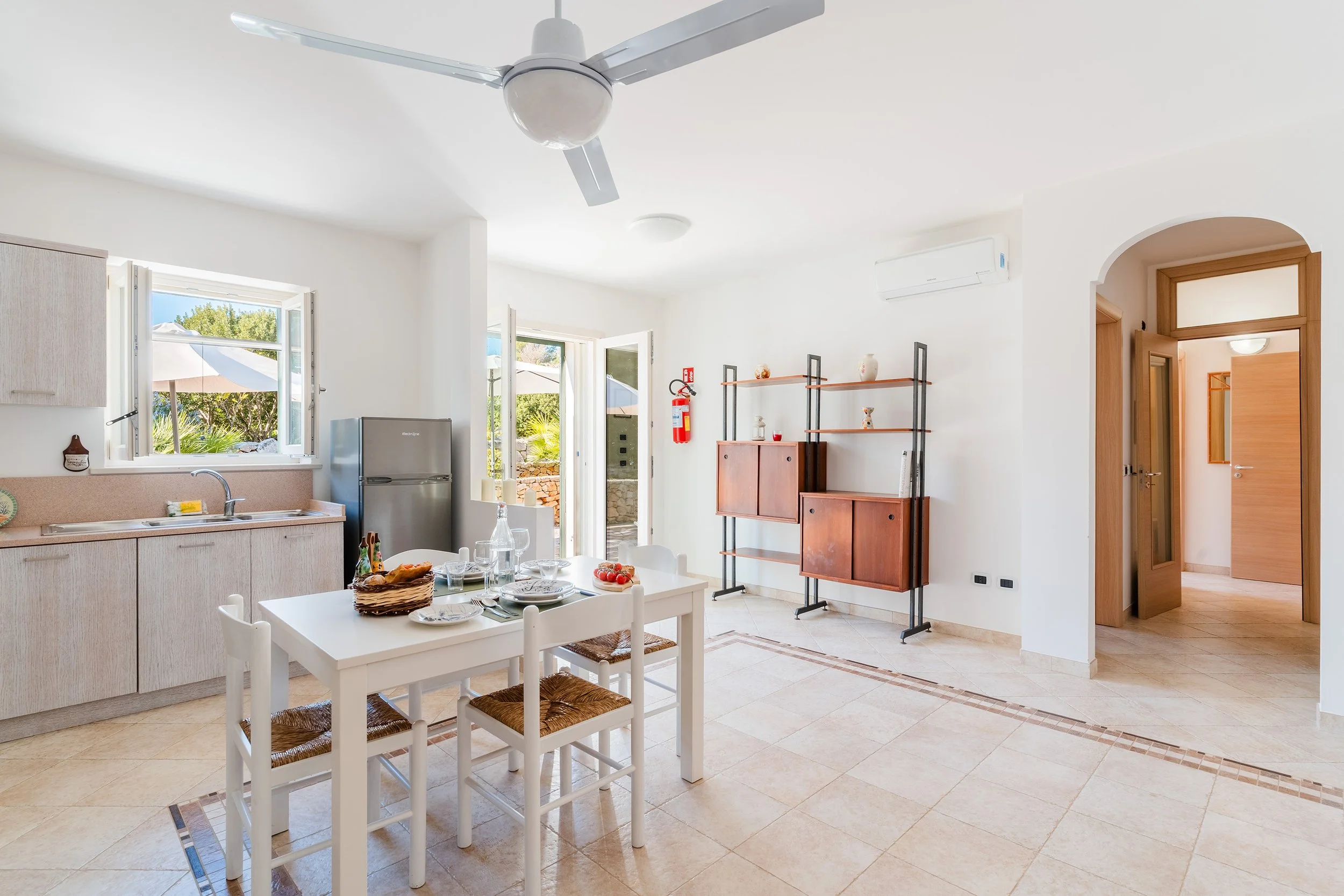 Bright, airy kitchen and dining area with white walls, open windows, and a ceiling fan. There is a white dining table with four chairs, decorated with a basket of bread, glasses, and plates. The kitchen has light wood cabinets, a small gray refrigera