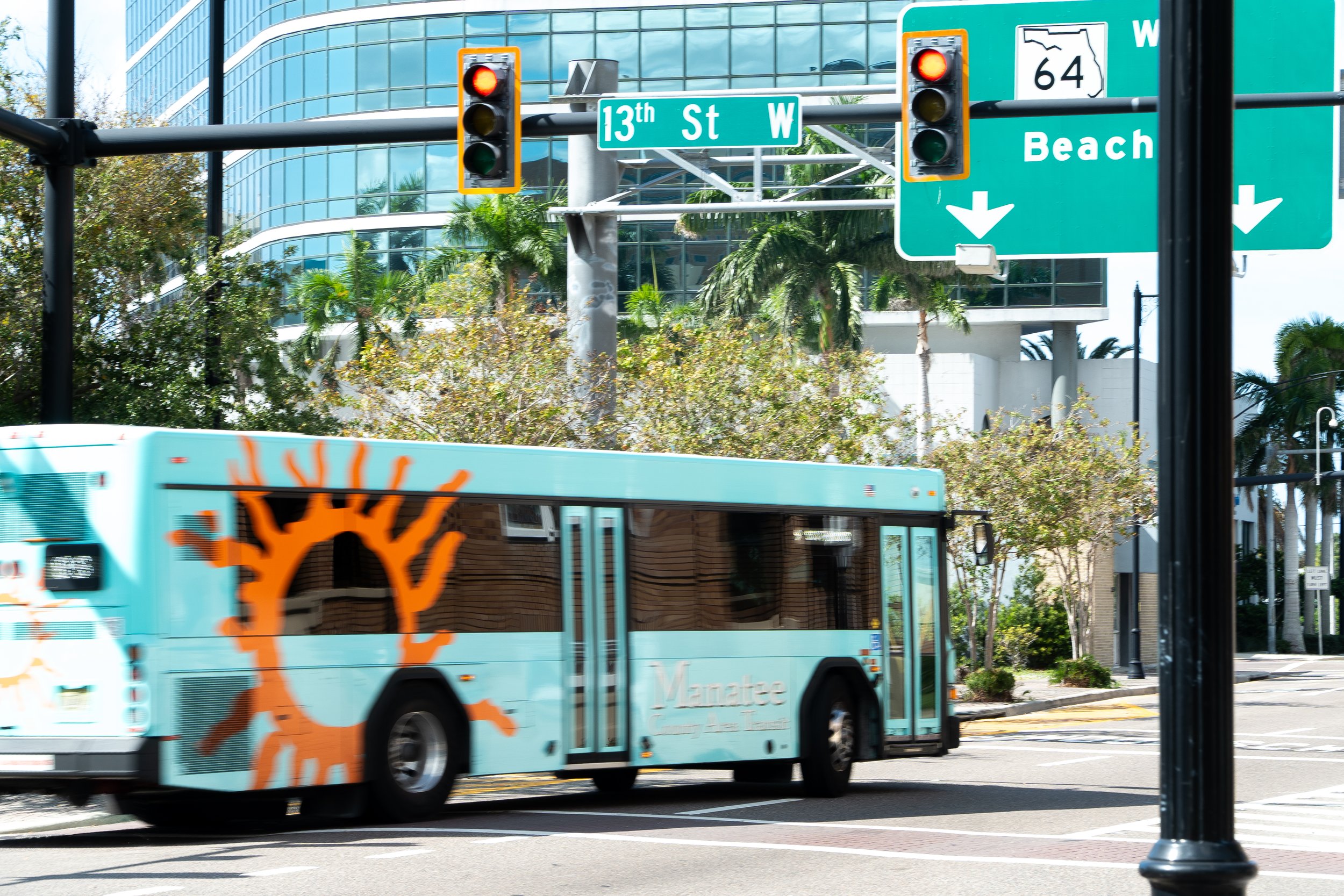 City street scene with a green street sign for 13th St W, a traffic light with red light on, a teal bus with an orange tree logo and the text 'Monatze' on its side, and modern glass buildings in the background.