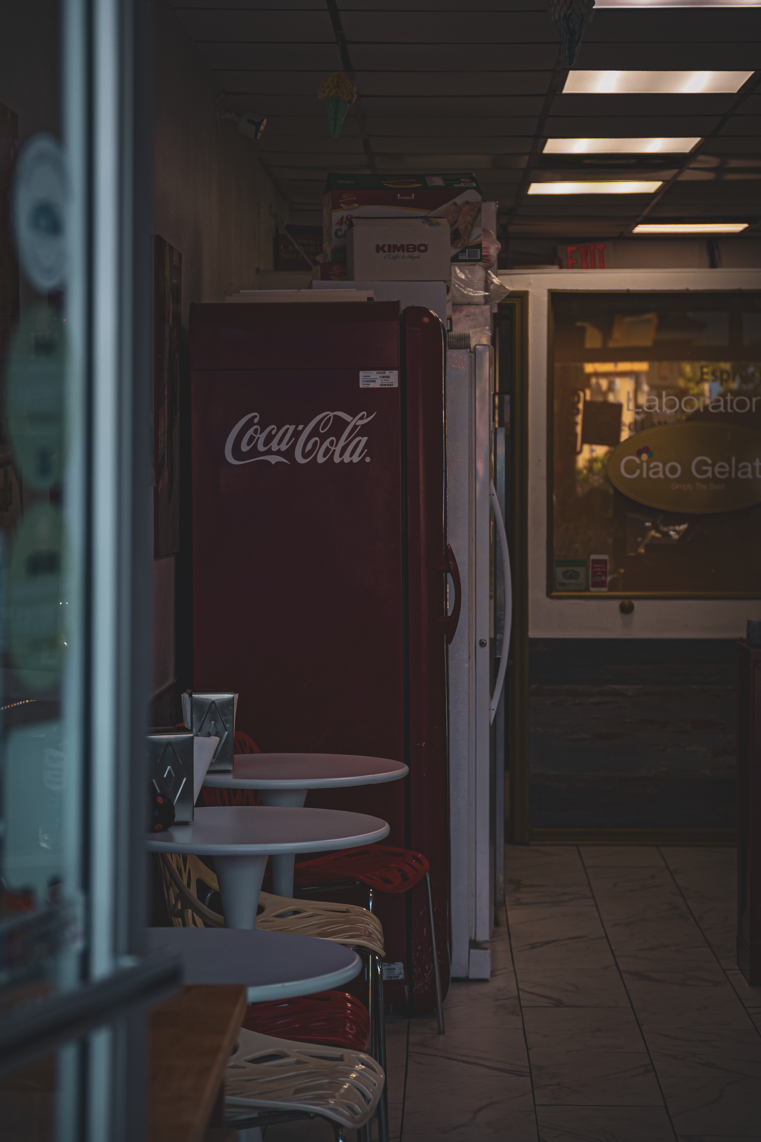 Inside a small eatery or convenience store with soda and snack machines, with a row of white and red chairs along the wall, and an exit door in the background.