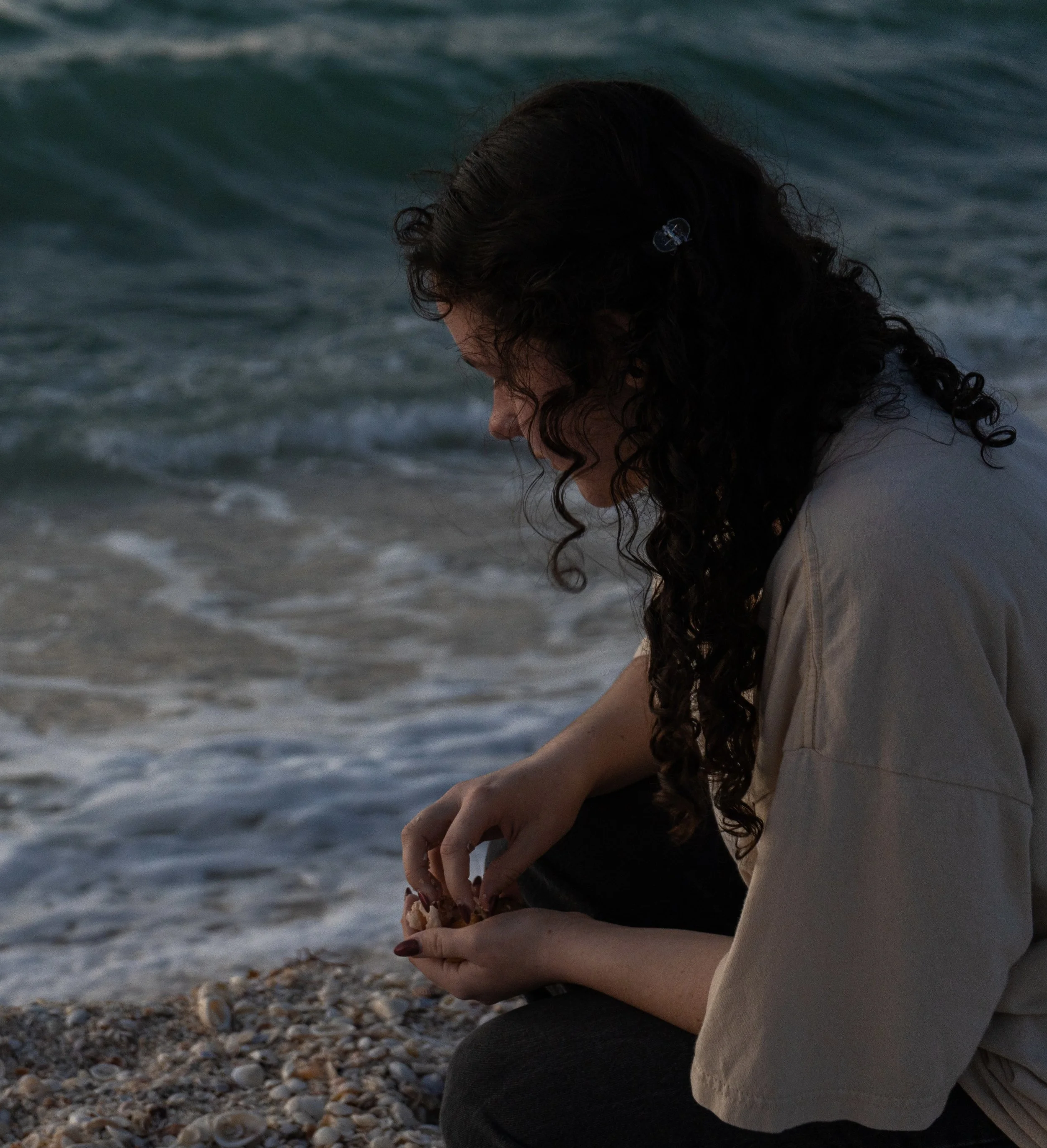 A woman with long, curly dark hair sits on a shell-covered beach near the ocean, holding a seashell in her hands. She is wearing a beige or light gray top and dark pants, leaning forward with her head bowed as she looks at the shell. The ocean waves are visible in the background.