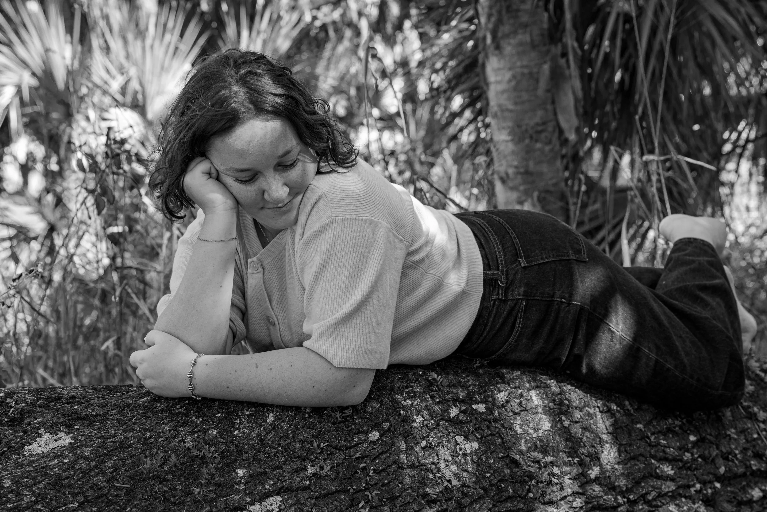 A woman with curly hair lying on her side on a large rock, resting her head on her hand, outdoors with trees and foliage in the background.