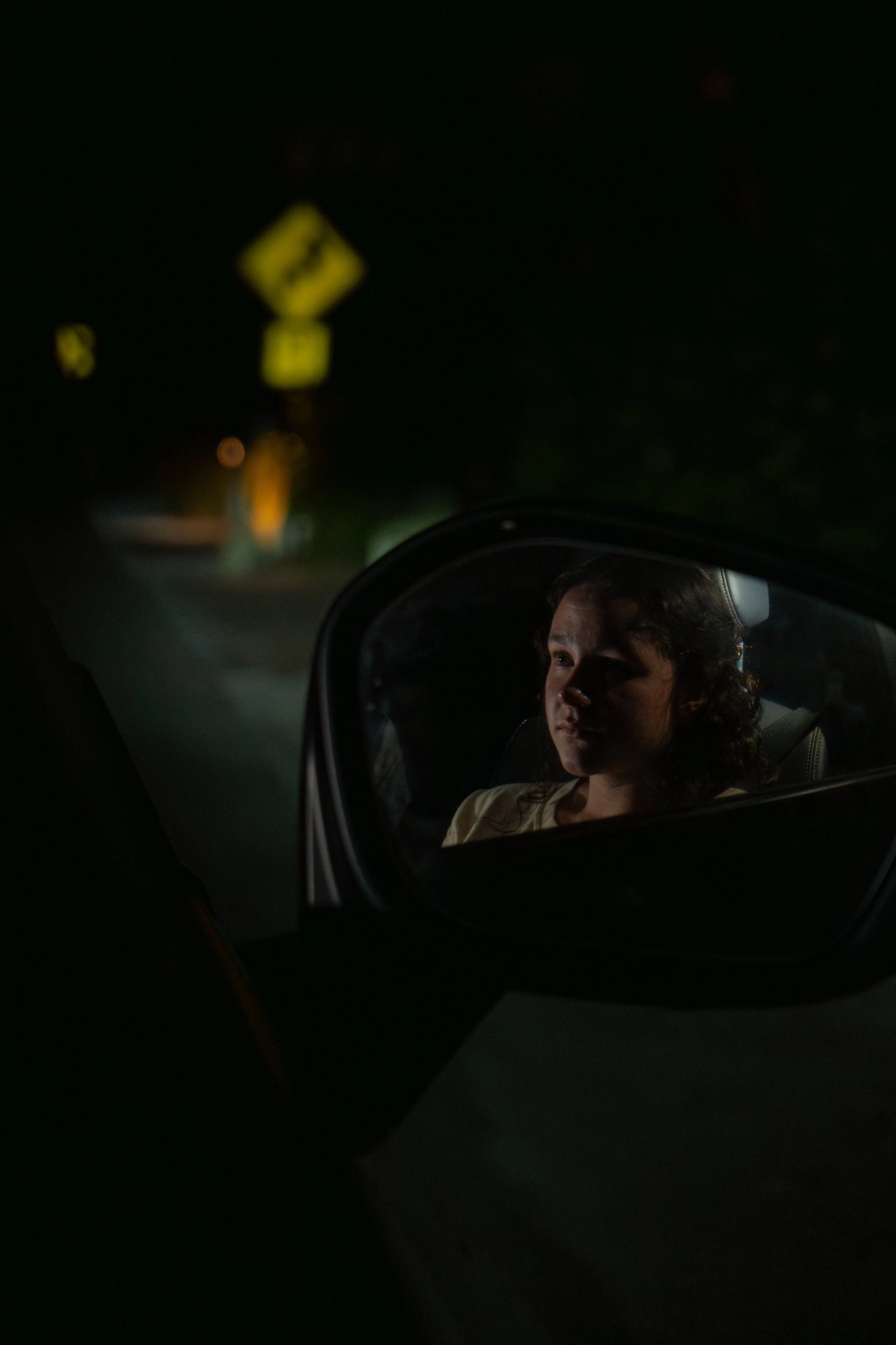 A woman with curly hair looking into the side mirror of a car at night, with a blurry yellow road sign in the background.