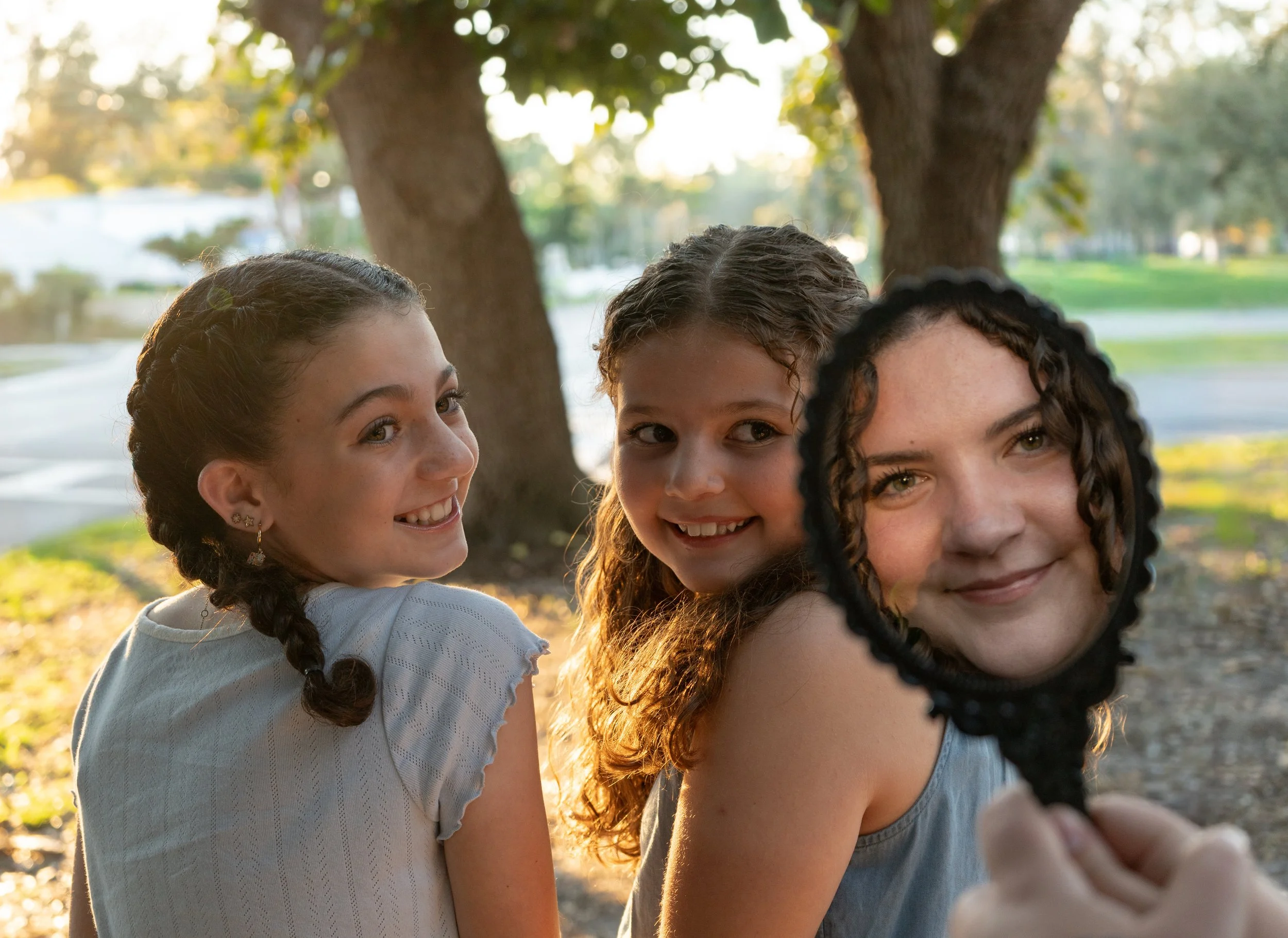 Three girls standing outdoors in a park, smiling, as sunlight filters through trees behind them.