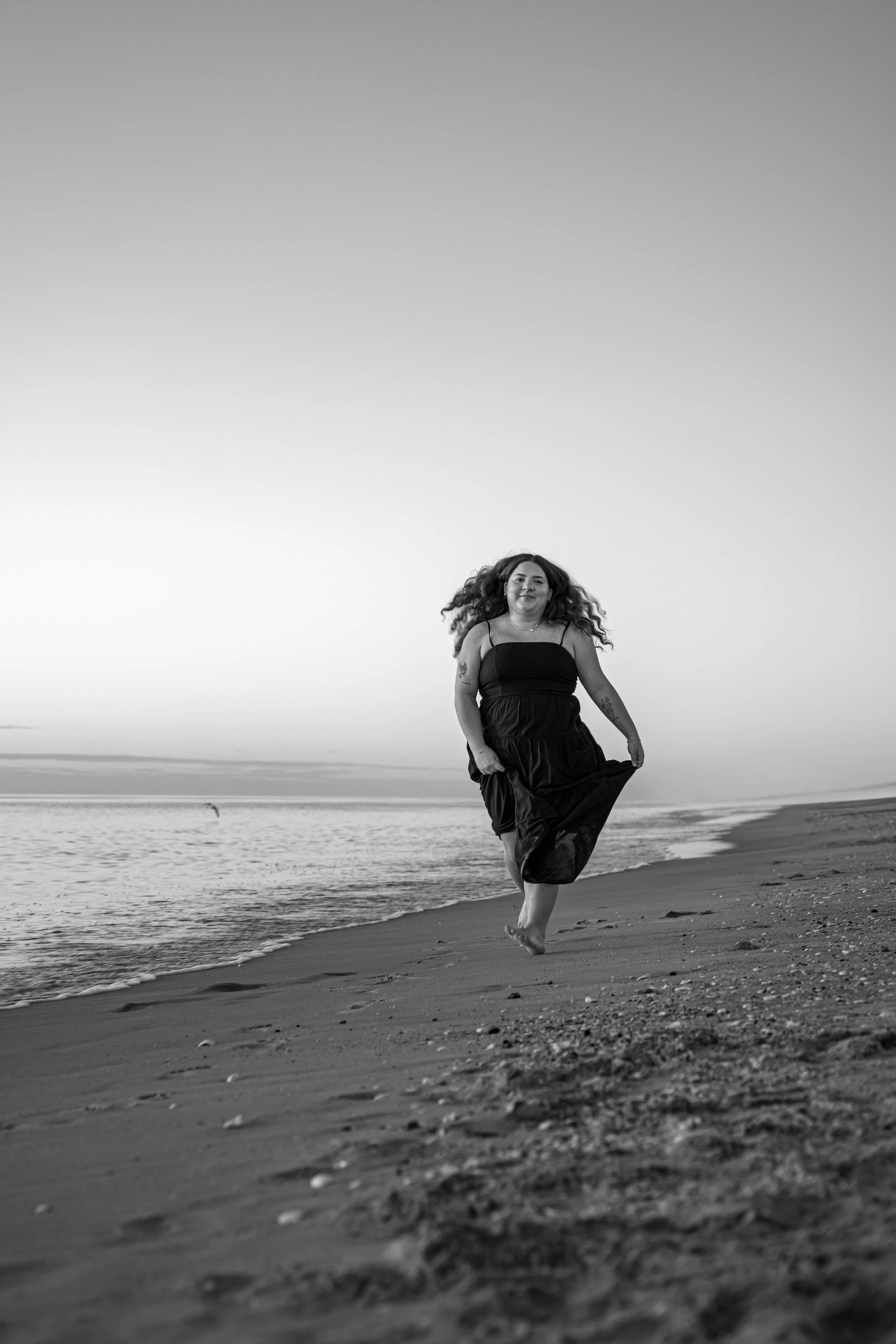 A woman with curly hair walking barefoot on the beach at sunset, holding her long black dress as she walks along the shoreline, with calm water and a clear sky in the background.