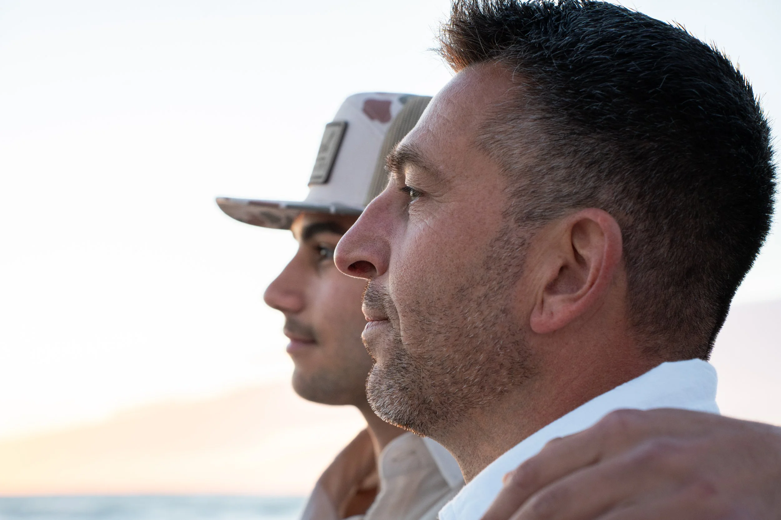 Close-up of a man and a woman standing side by side outdoors during sunset, looking out towards the horizon with calm and serene expressions.
