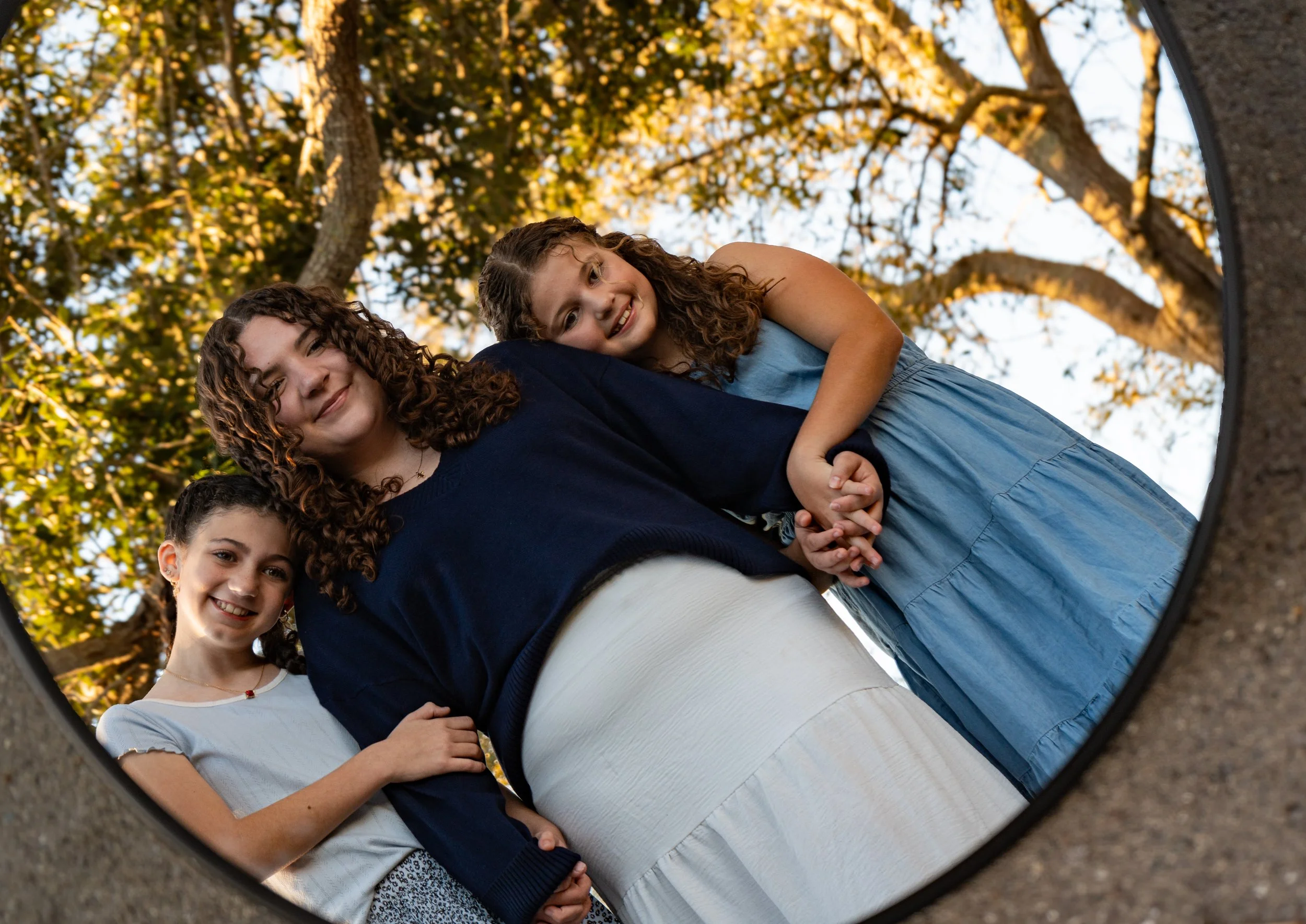 A group of smiling children and a woman are seen in a mirror reflection outdoors, with trees and blue sky in the background.