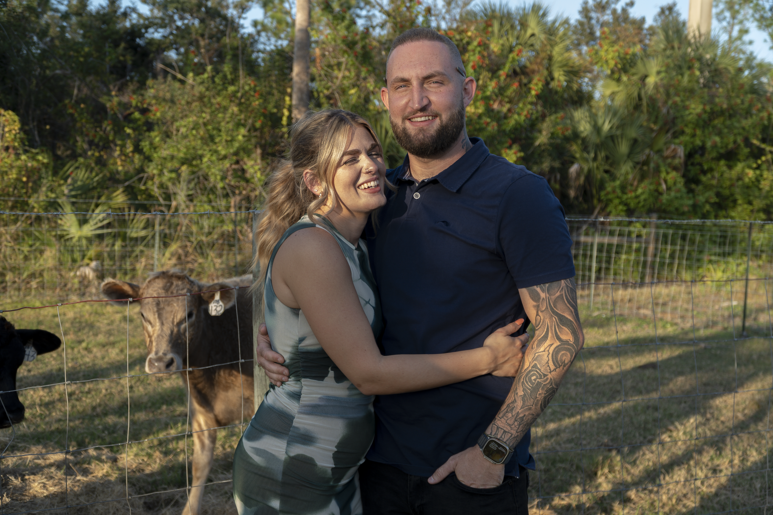 A smiling man with a beard and tattooed arm hugging a woman with wavy hair, standing in front of a fence with cows and lush green trees in the background.