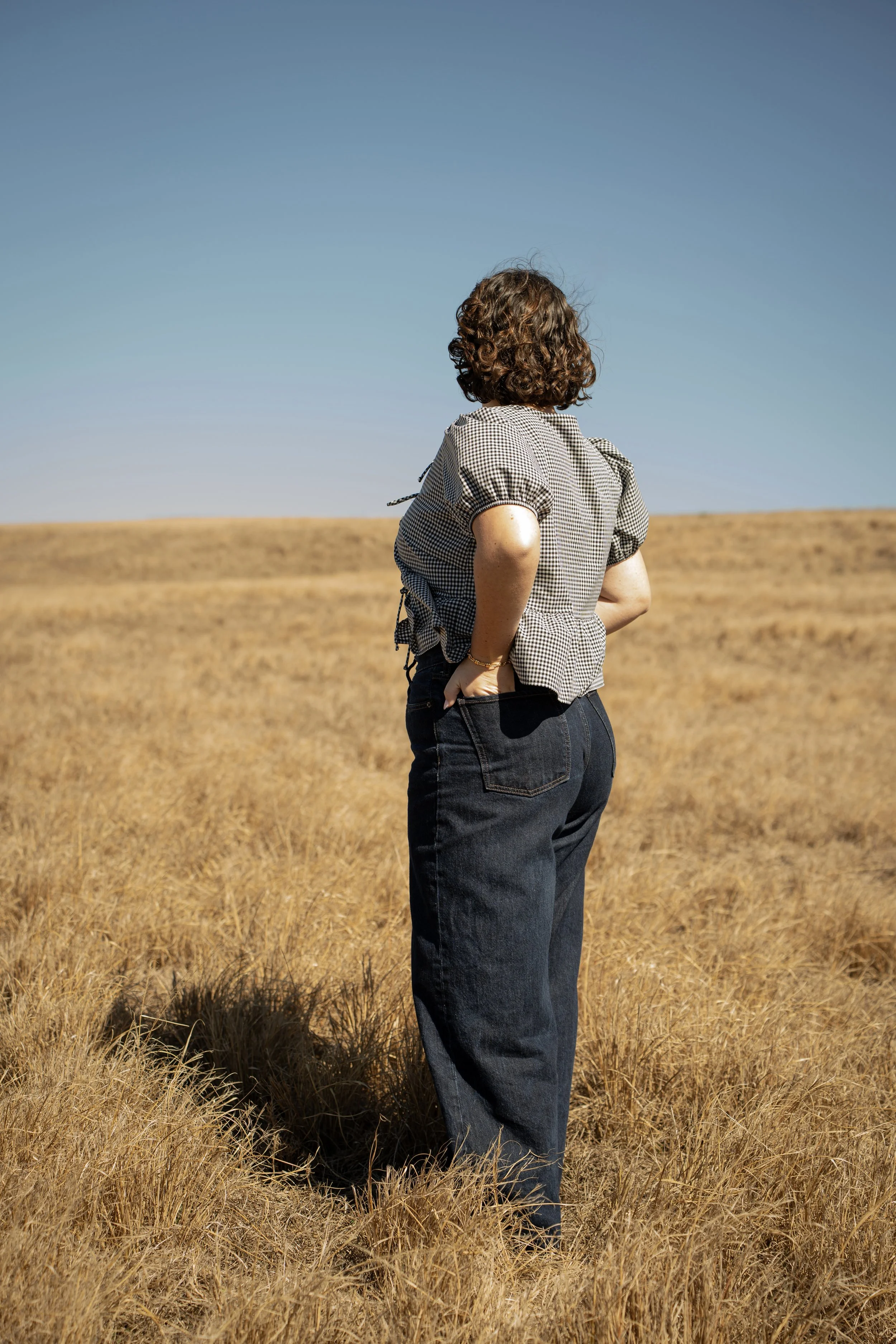 A woman with curly hair stands in a field of tall, dry grass, facing away from the camera, under a clear blue sky.
