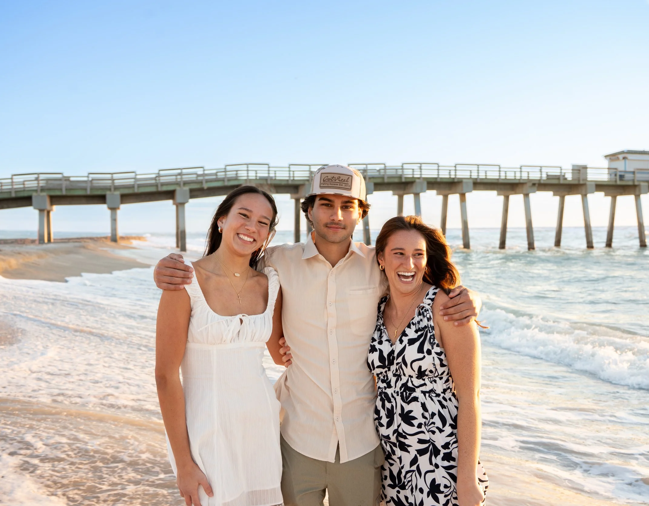 Four people standing close together on the beach in front of a pier, smiling and embracing each other, with blue sky and ocean in the background.