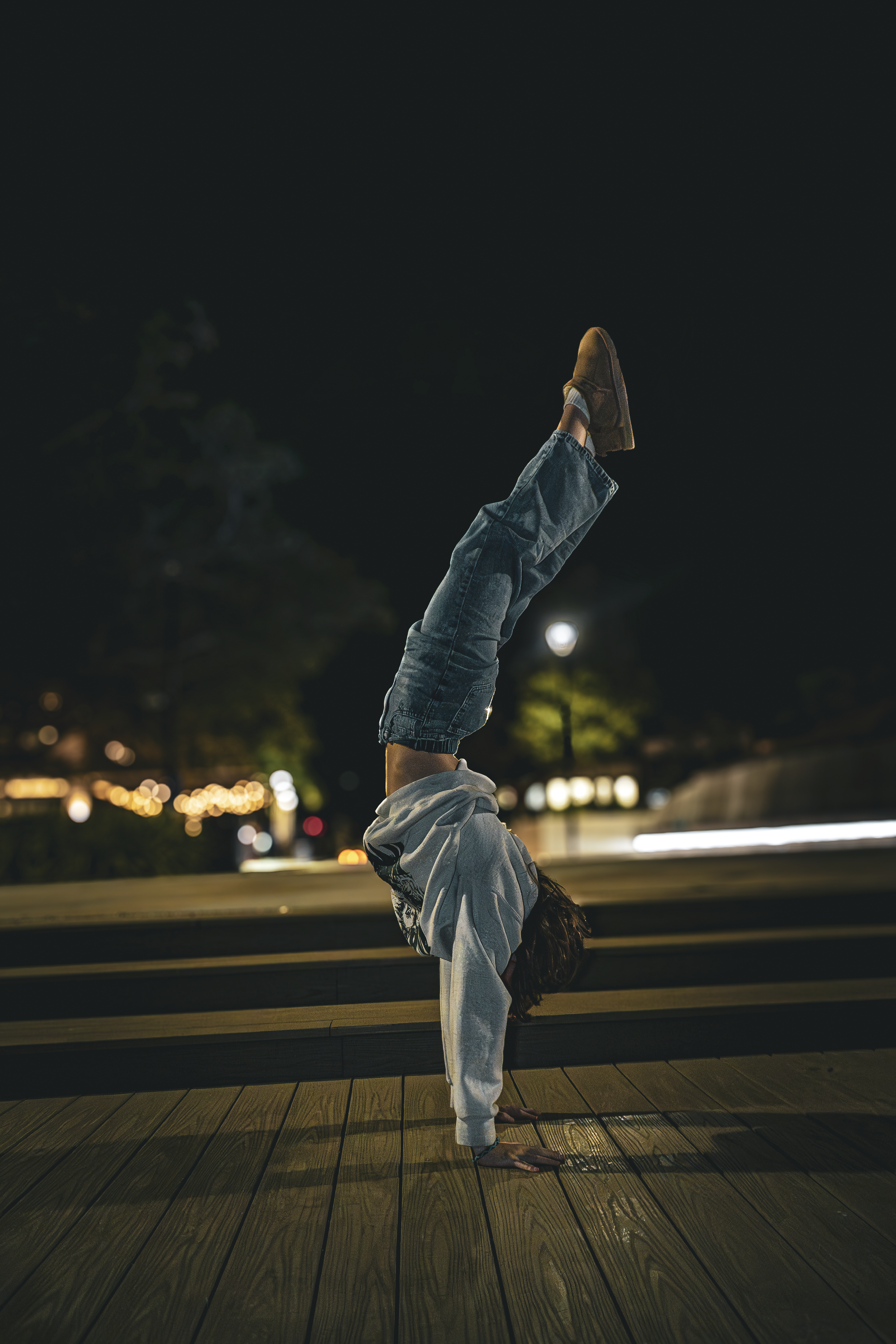 A person doing a handstand at night on a wooden deck in an urban park, with city lights and trees in the background.