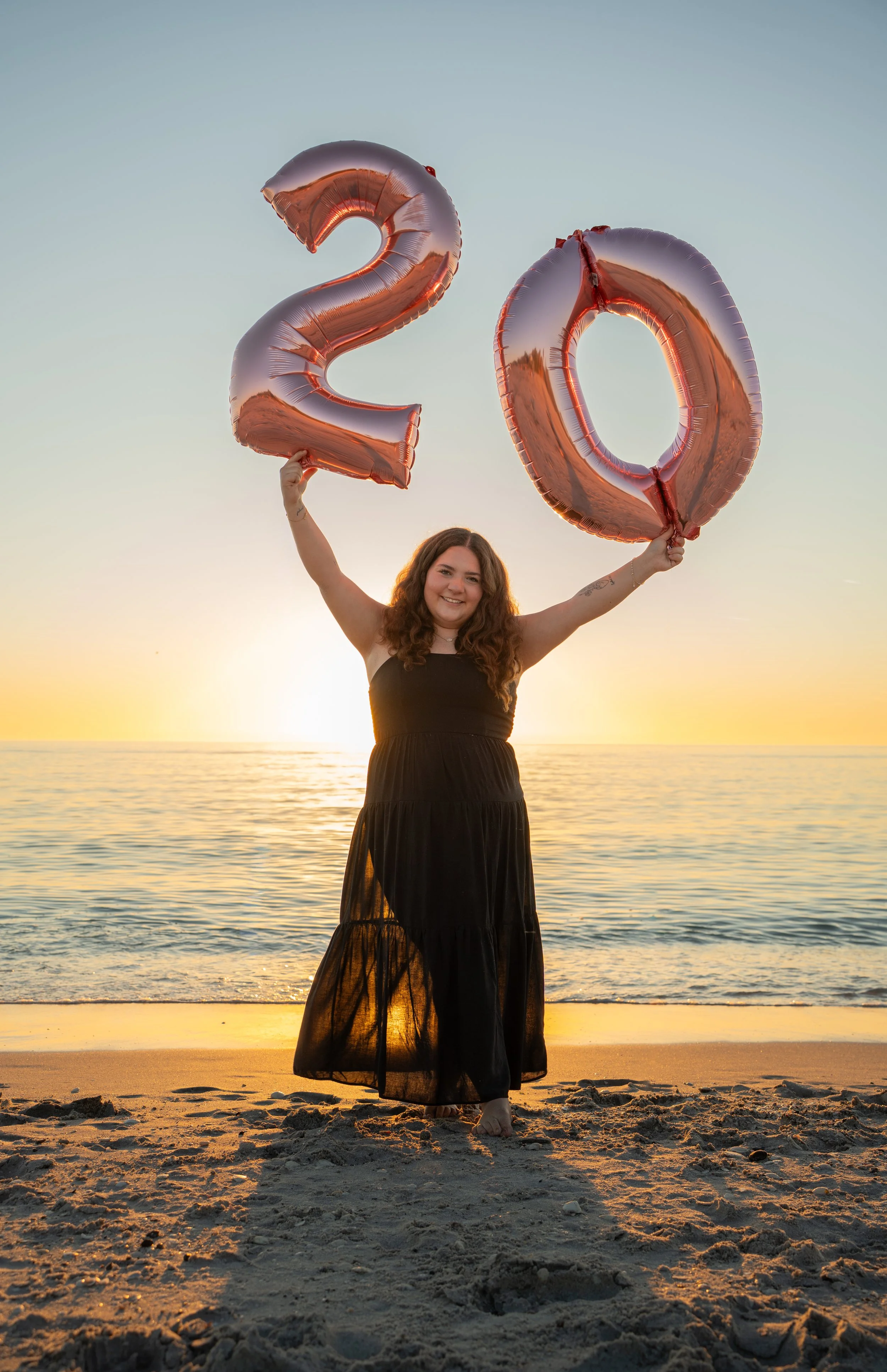 A woman standing on a beach at sunset holding large rose gold foil balloons shaped as the numbers 2 and 0, celebrating a 2020 event.