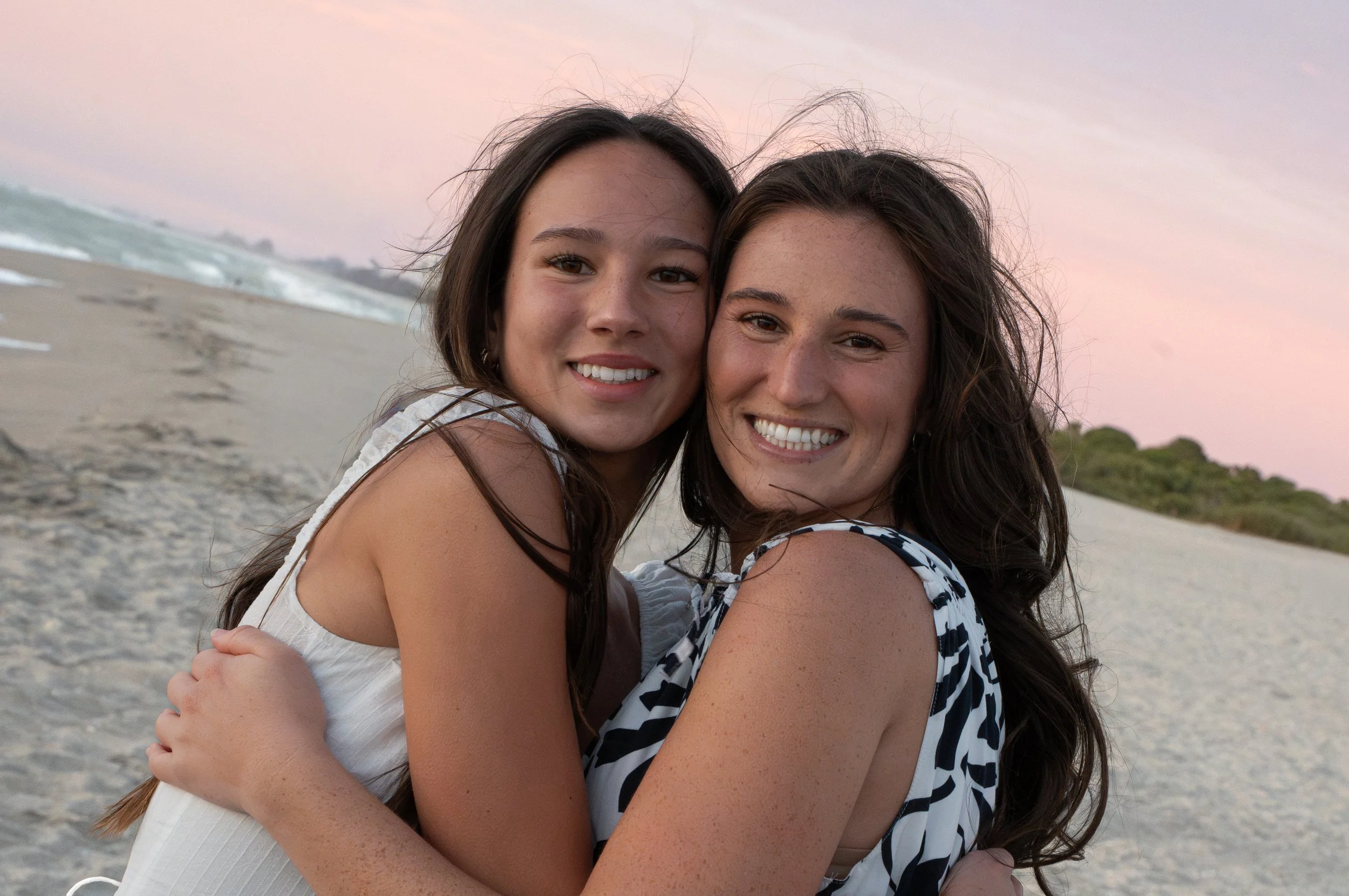 Two young women hugging and smiling on a beach during sunset, with the ocean and sandy shore in the background.