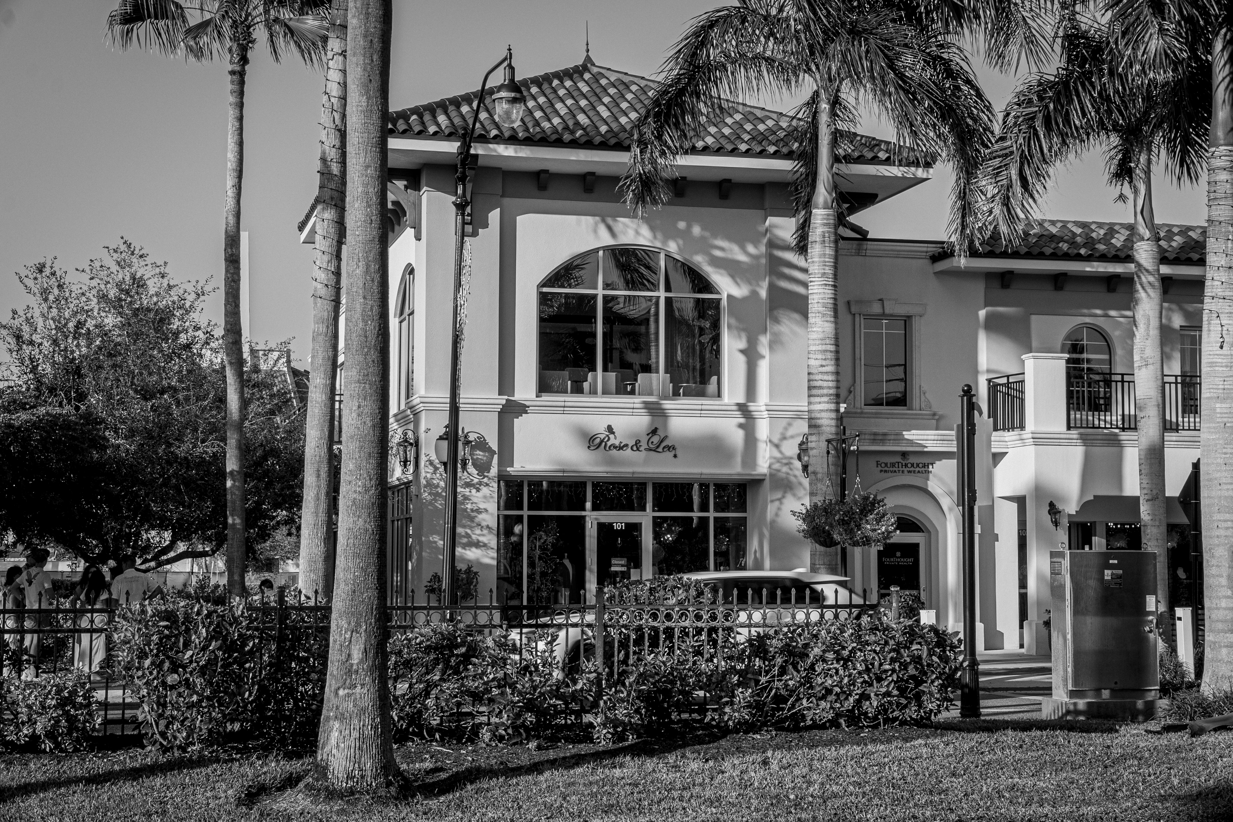 A black and white photo of a multi-story building with a terracotta tile roof, large arched windows, and a sign that reads 'Rose & Lea'. There are tall palm trees in front of the building and a streetlamp on the sidewalk. Few people are visible in th
