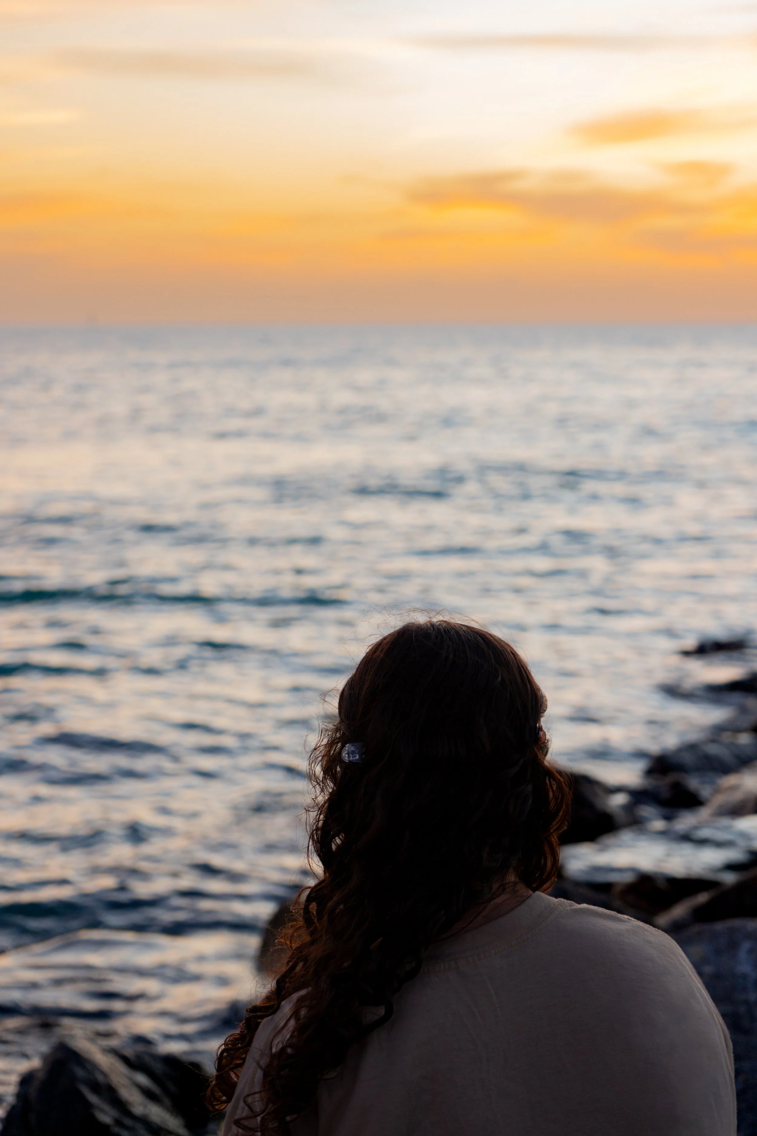 A person with long dark curly hair and glasses looking out over the ocean at sunset, with orange and yellow clouds in the sky.