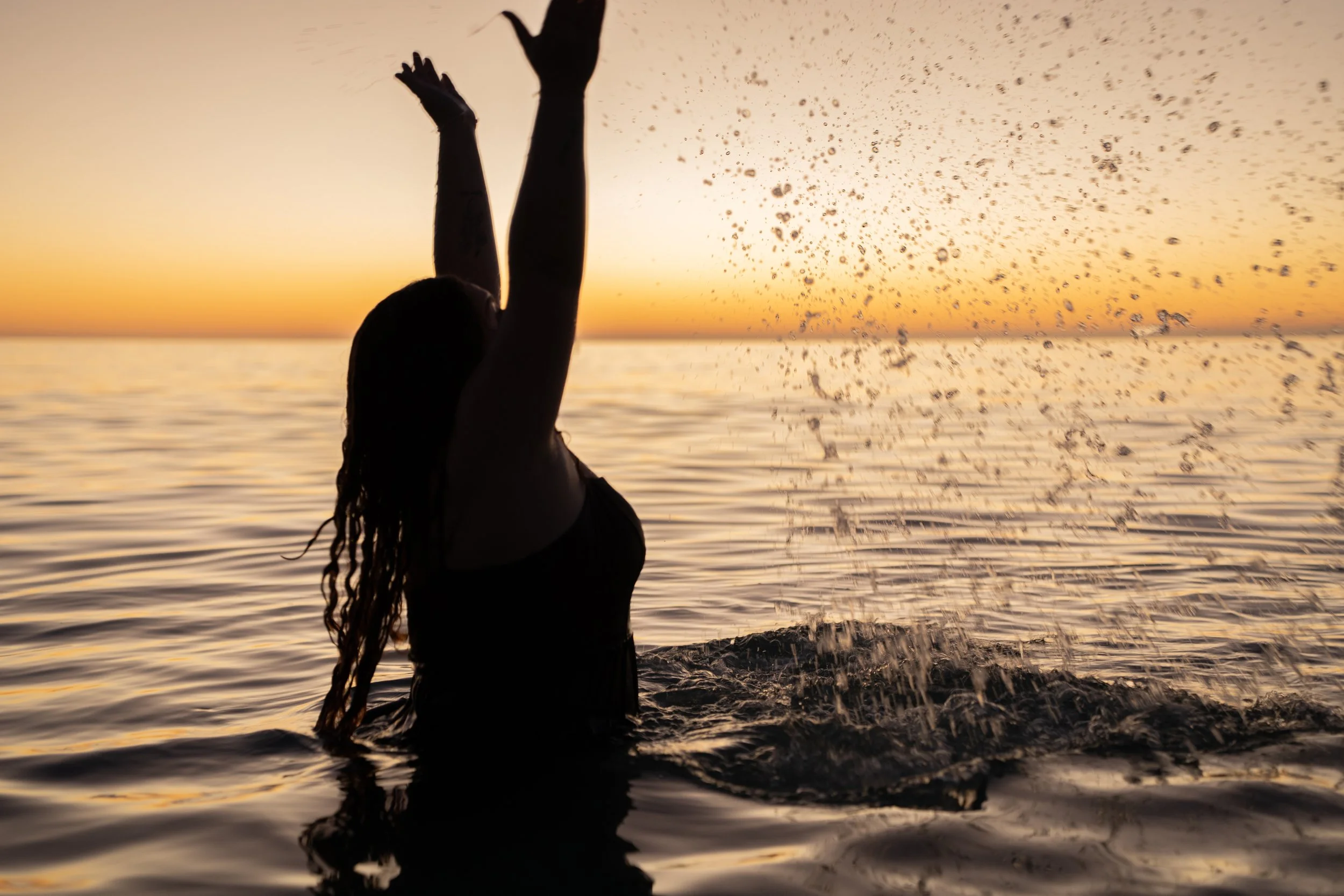 A silhouette of a person splashing water in the ocean during sunset.