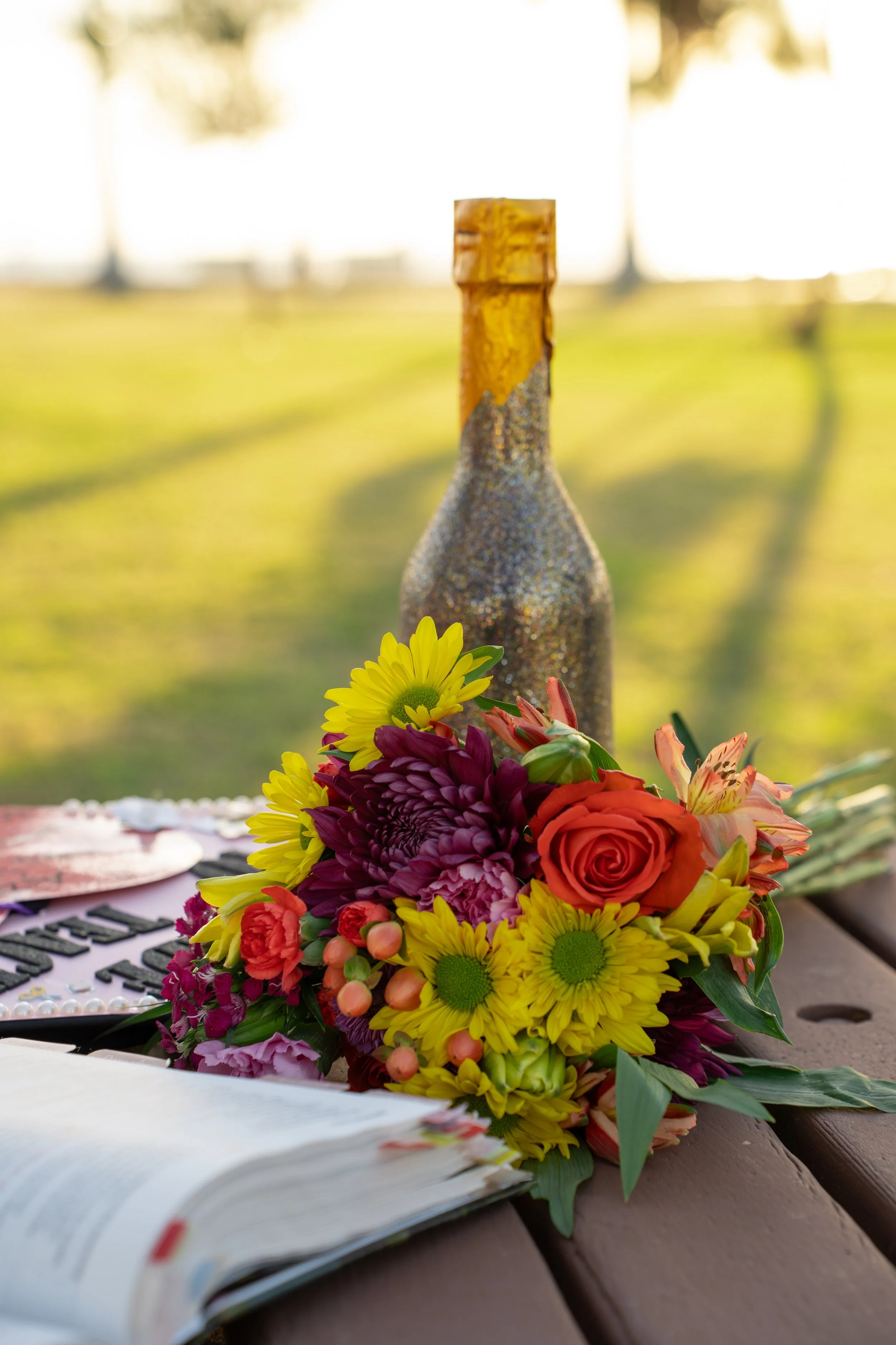 A colorful bouquet of flowers, including yellow daisies, purple and pink flowers, with a wine bottle in the background, on a wooden surface outdoors.