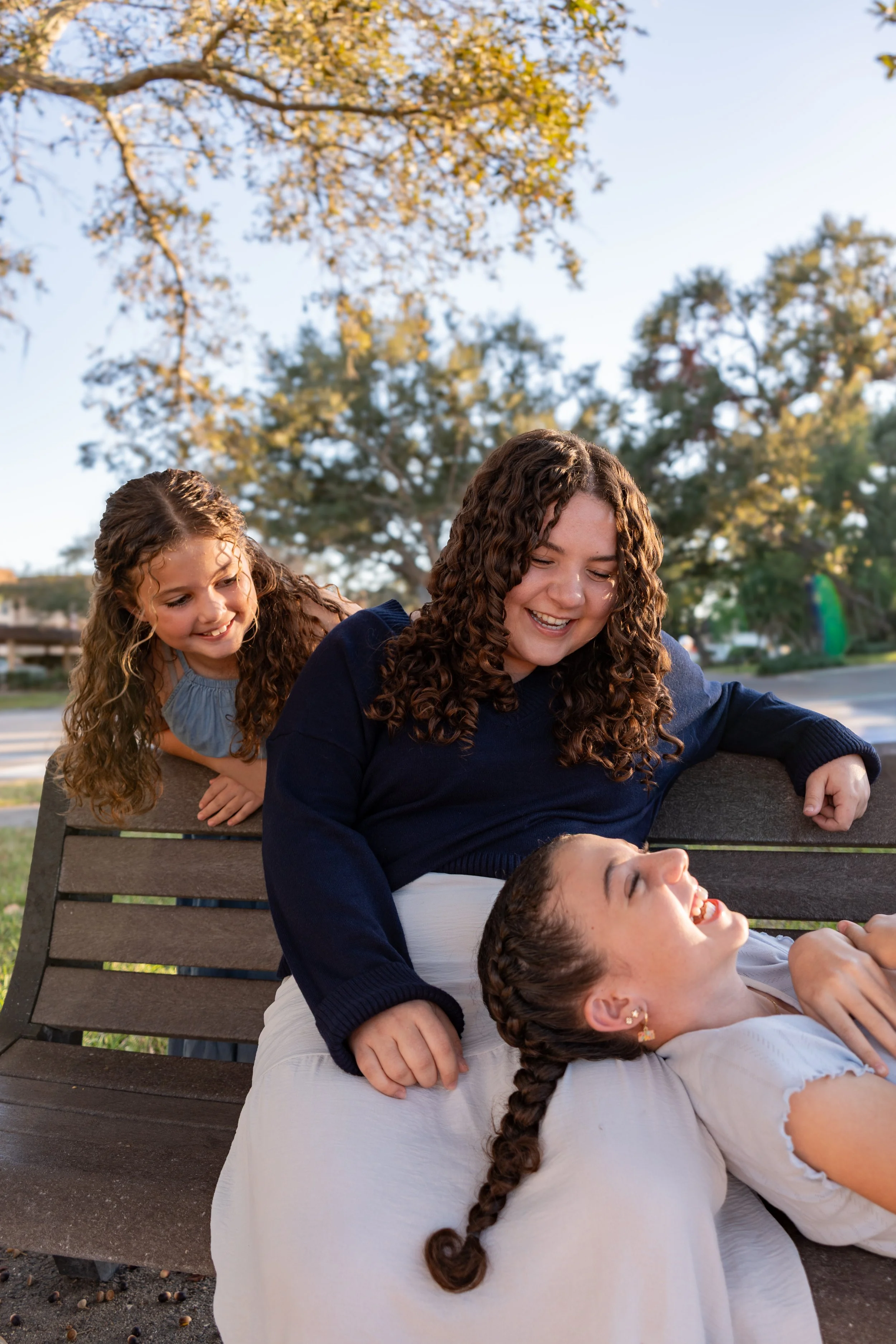Four people, including two young girls and two young women with curly hair, are sitting and lying on a park bench outdoors during the daytime. They are smiling and laughing, engaged in a joyful moment. The background features trees with green and yellow leaves and a clear sky.