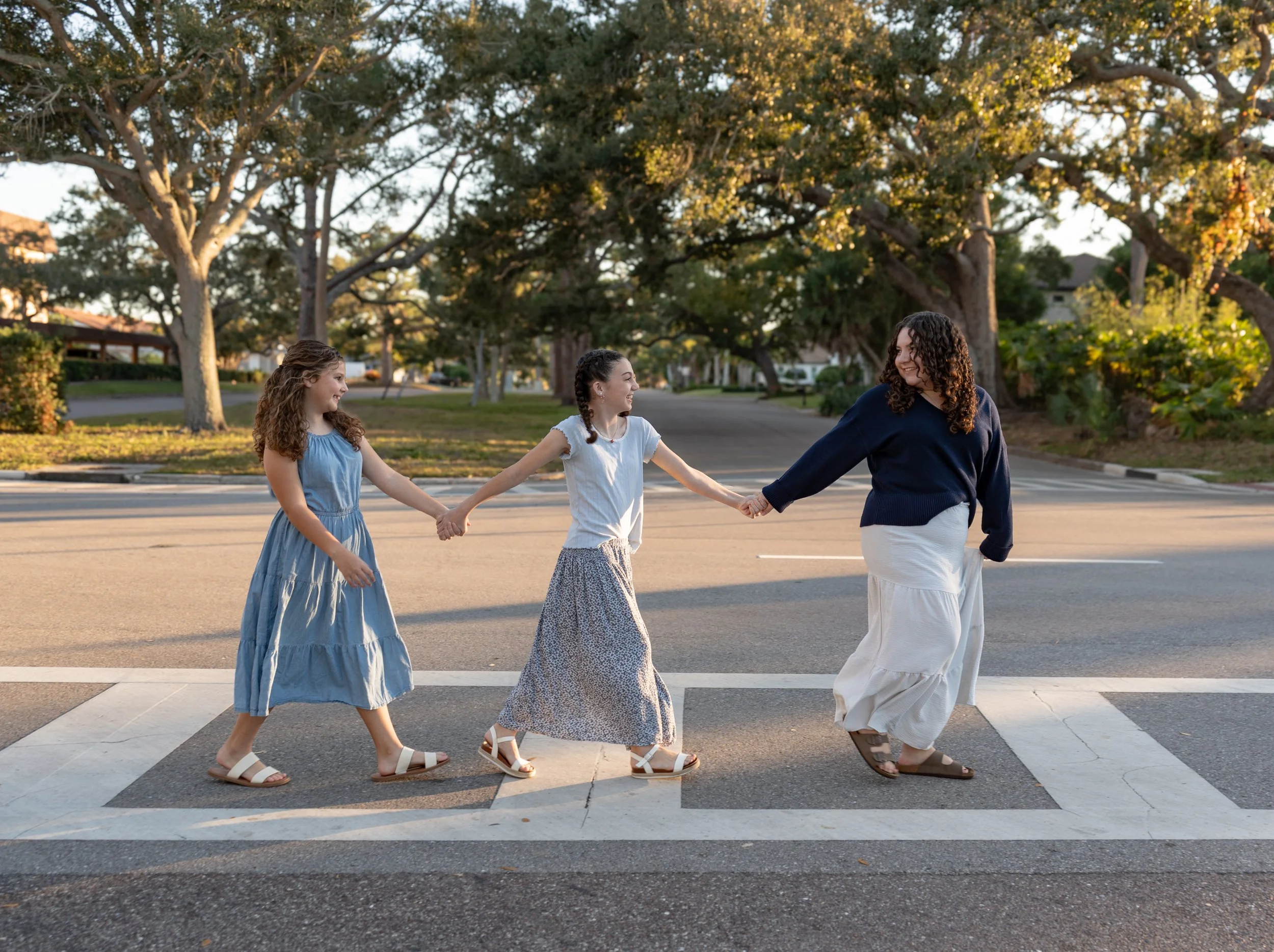 Three women holding hands and walking across a crosswalk in a neighborhood park with large trees and houses in the background during the daytime.