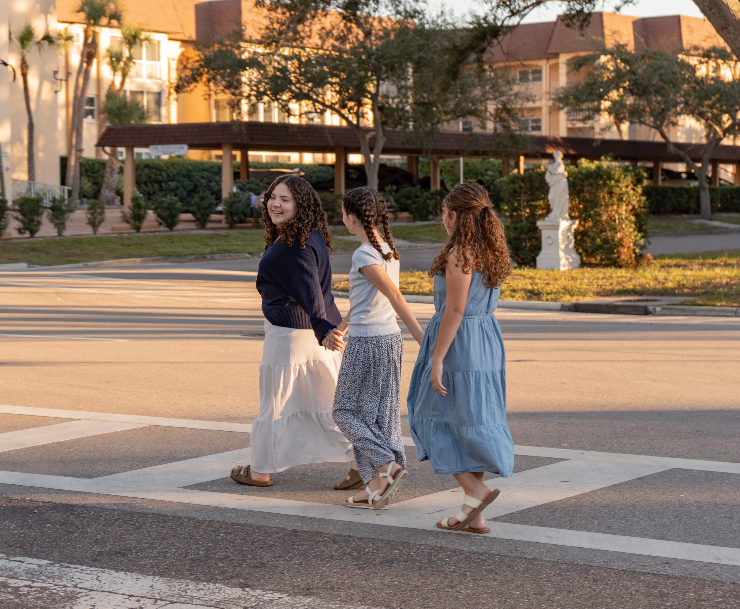 Three girls holding hands and walking across a crosswalk in a neighborhood during sunset, smiling and enjoying each other's company.