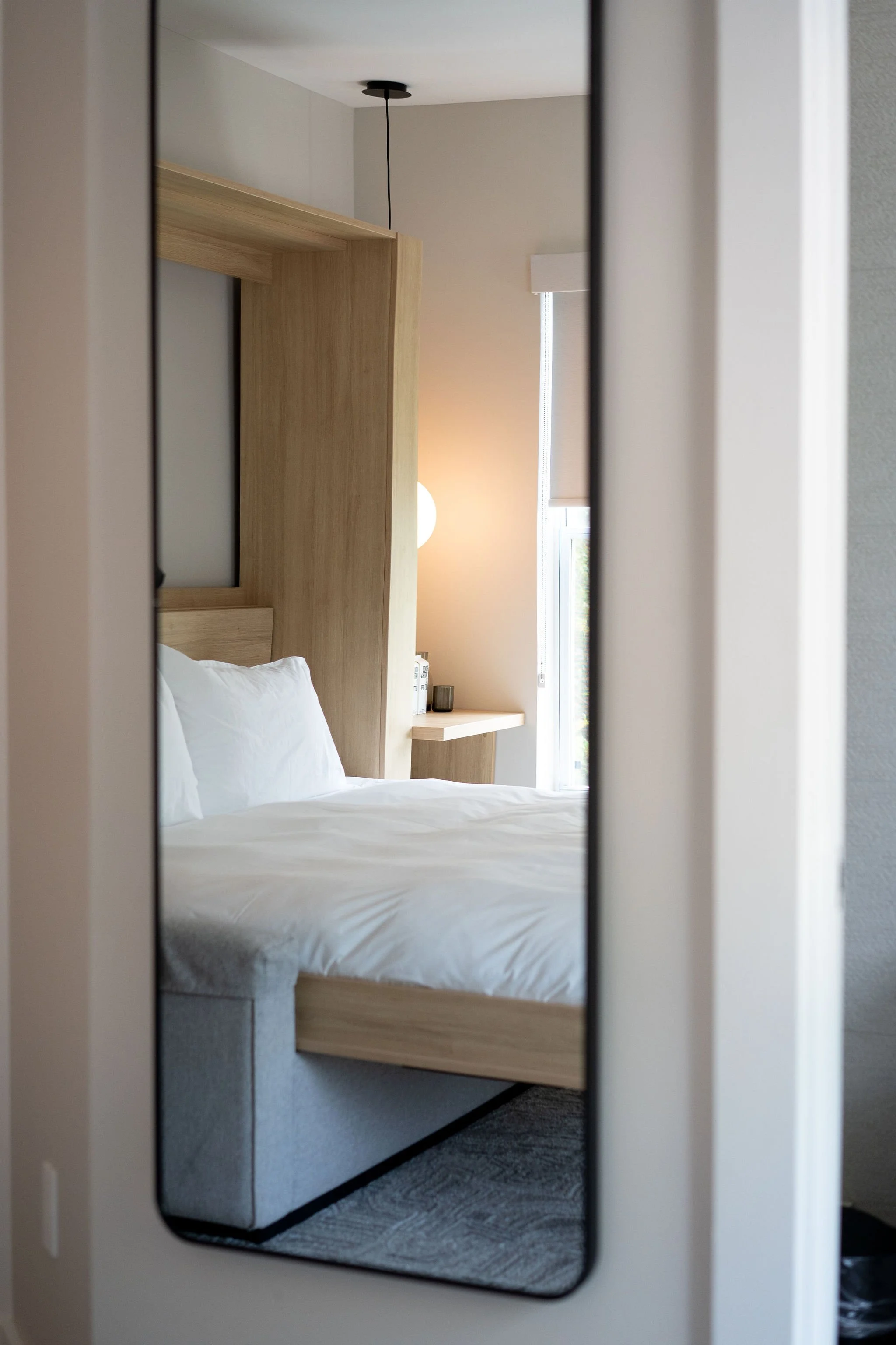 A reflection of a hotel room with a white bed, wooden headboard, and a small side table near a window, seen in a mirror.