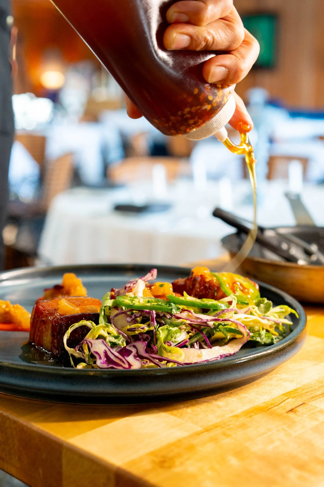 A hand squeezing a bottle of barbecue sauce onto a plate of food, which includes grilled meat and a salad with shredded cabbage and other vegetables, on a wooden table in a restaurant setting.