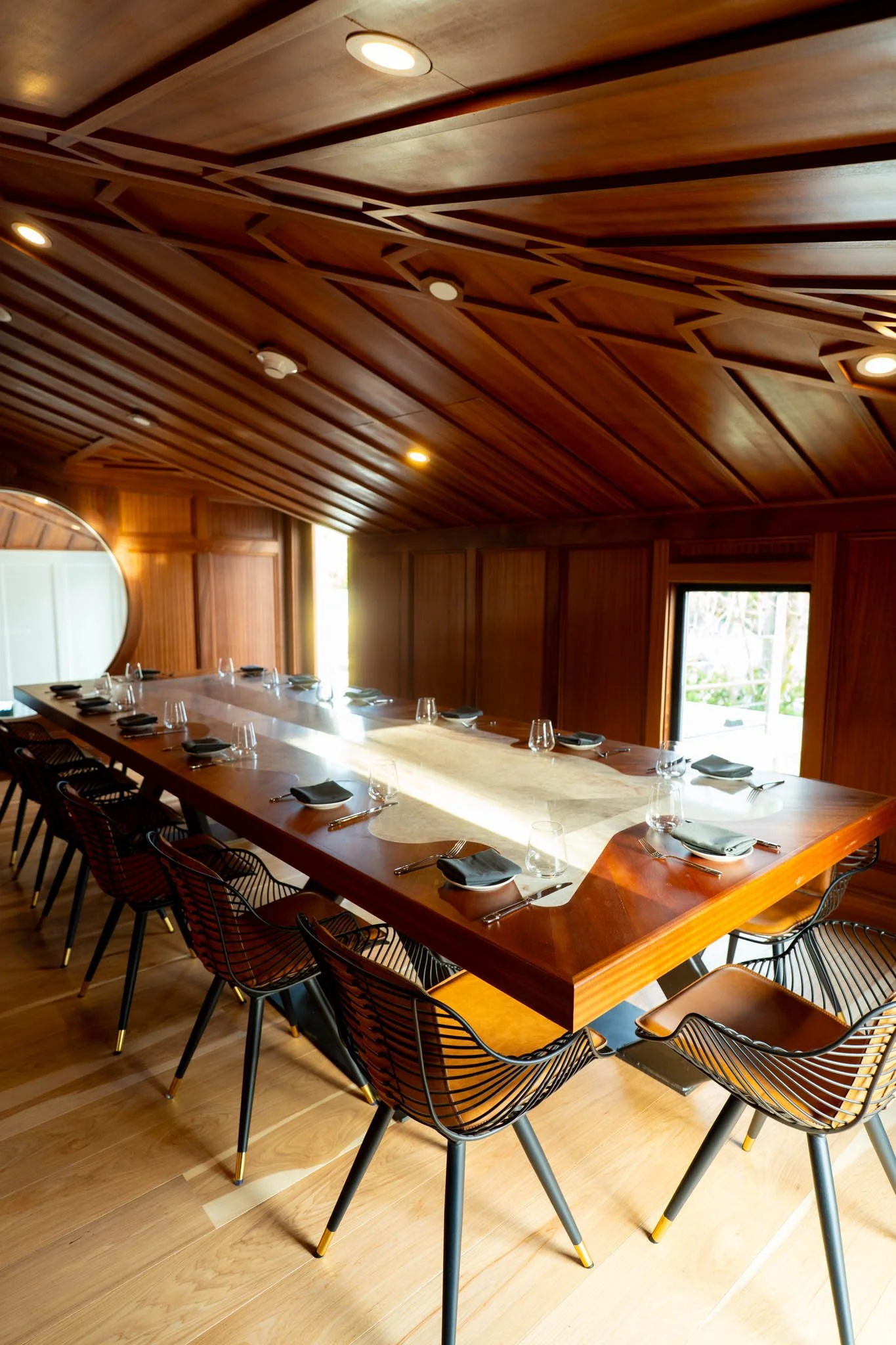 A modern dining room with a large wooden table set with black napkins, plates, and glasses, surrounded by black wire chairs with wooden seats, featuring warm wood-paneled walls and ceiling, and windows letting in natural light.