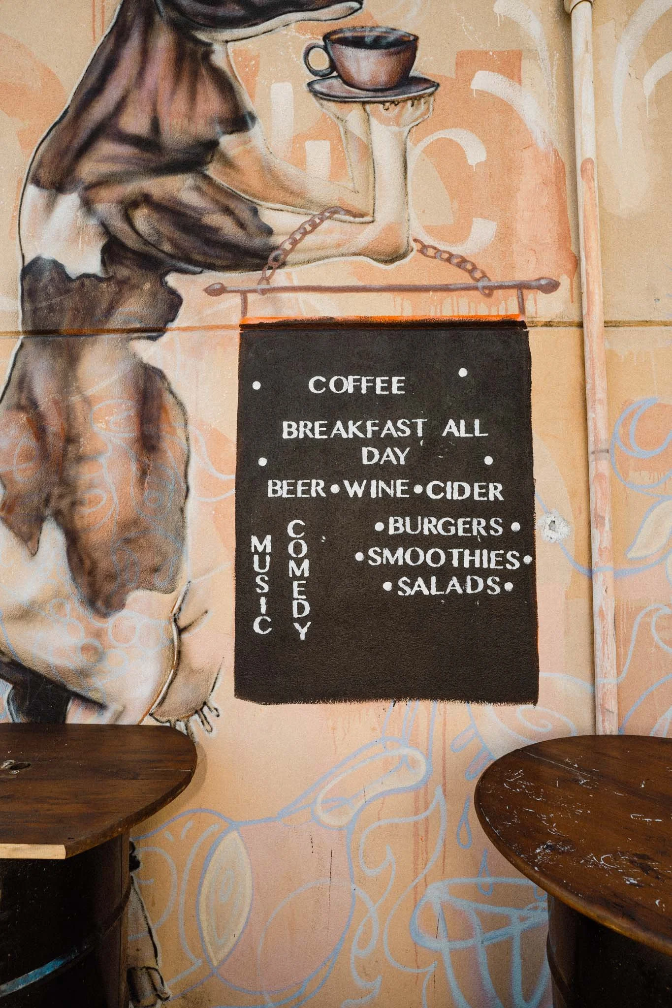 A mural of a person holding a tray with coffee cups. A blackboard menu lists food and drink items such as coffee, breakfast all day, beer, wine, cider, burgers, smoothies, and salads.
